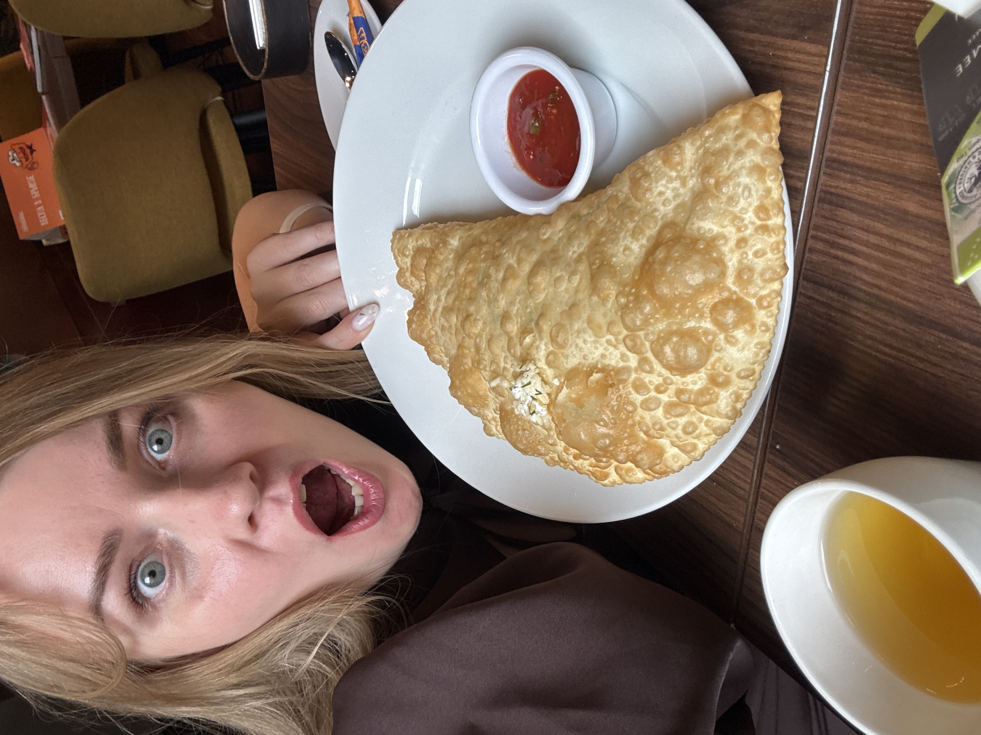 A young woman with blonde hair and blue eyes is pictured at a table. Her mouth is open wide in excitement or surprise. In front of her is a white plate with a large, fried, triangular pastry, likely a cheburek, and a small white dish containing a red sauce. To the right of the plate is a white cup filled with a yellow liquid, possibly tea or juice. The table has a wood-grain surface. The location is likely a restaurant in Saint Petersburg, Russia, judging by the food served.