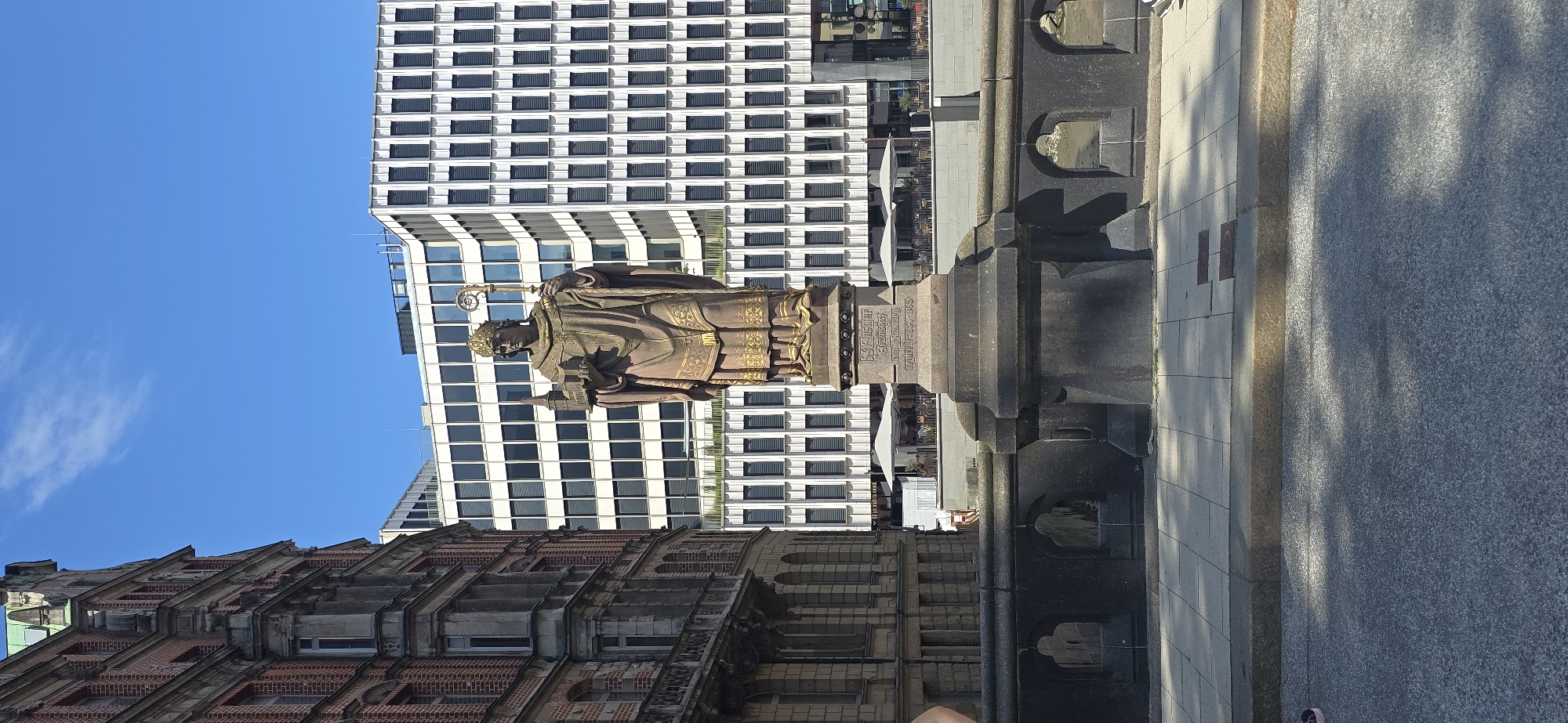 The image shows the Saint Ansgar statue in Hamburg, Germany, a bronze sculpture of a bishop holding a model of a cathedral in his left hand and a staff in his right. The statue is positioned in front of a modern white building with many windows, and to the left, there is an older brick building with ornate architecture. The statue stands on a stone pedestal with inscriptions. The surrounding area appears to be a public space with a walkway and a street. The sky is clear and blue.