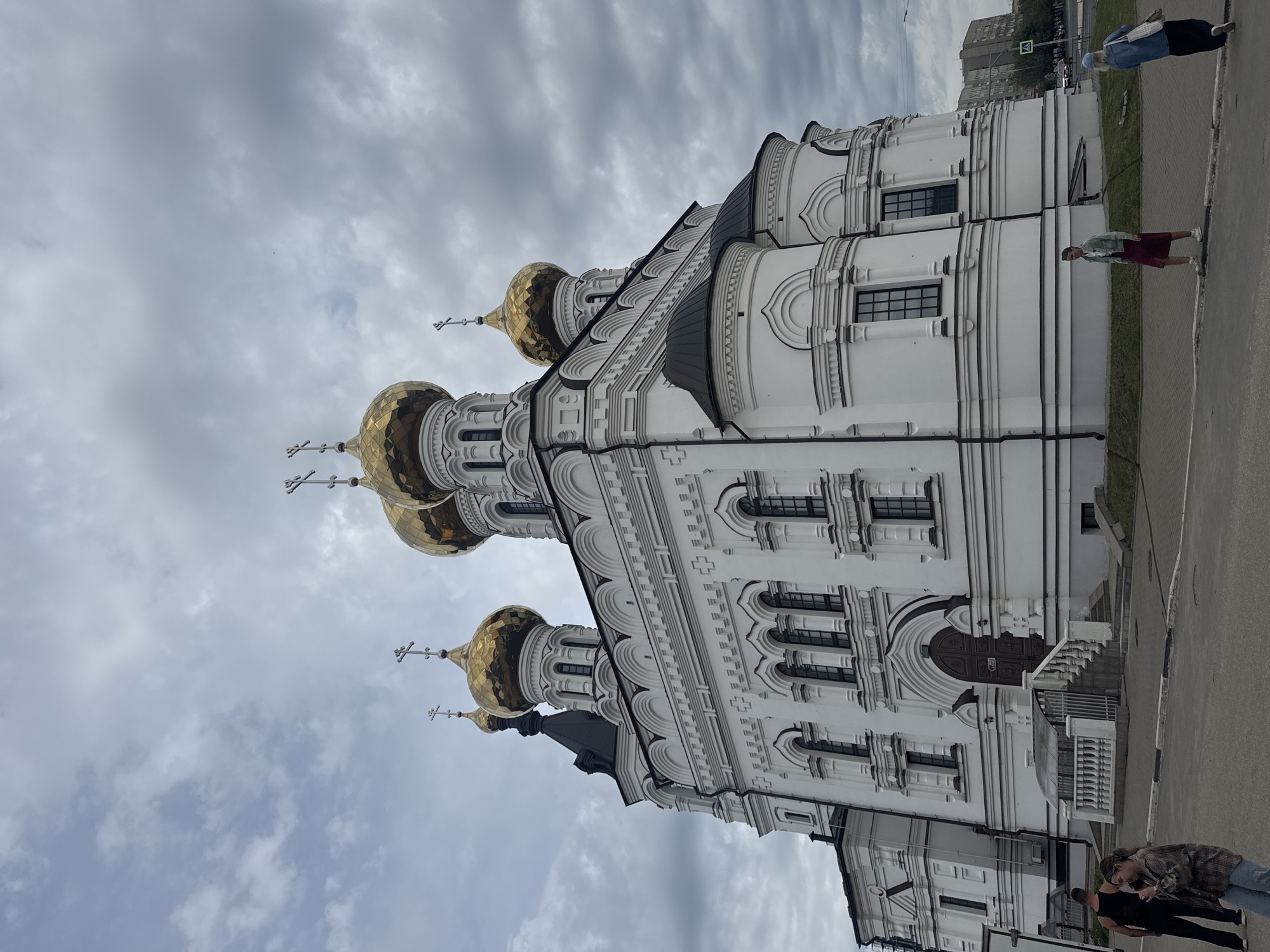 This is a low-angle, full-shot photograph of the Ascension Cathedral in Tver, Russia, under a cloudy sky. The cathedral is a large, white Orthodox church with multiple golden domes topped with crosses. It features ornate white architectural details, arched windows, and a dark-tiled roof. To the left of the cathedral is a smaller white building.
In the foreground, a paved area with a sidewalk separates the viewer from the cathedral. Several people are present:
* On the left, a man in a black tank top and black pants stands near a woman in a plaid shirt and jeans. They are looking away from the camera.
* In the middle ground, a woman in a light green jacket and a burgundy skirt walks across the paved area.
* On the right, a woman in a denim jacket and a dark skirt walks away from the camera, carrying a bag.
In the background, there is a street with a few cars and some apartment buildings. The overall scene suggests an overcast day in an urban environment.