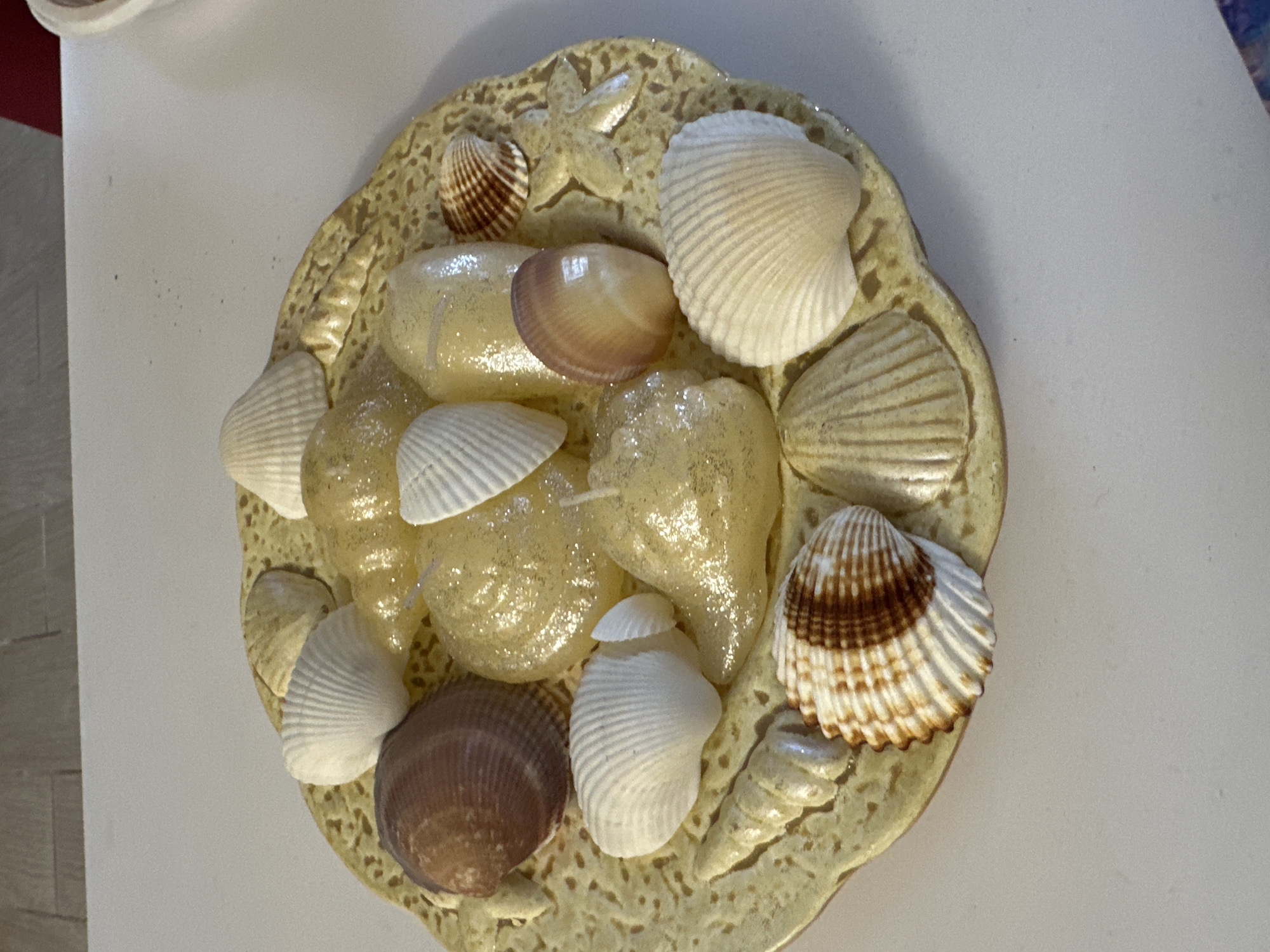 The image shows a decorative plate filled with seashells and glass beads. The plate has a beige color with a textured surface resembling sand or stucco, decorated with molded shells and a starfish. The seashells on the plate are of various sizes and colors, including white, brown, and striped patterns. Glass beads are interspersed between the shells. The background is a white surface. There is no information about the location in the city of Sofia, Bulgaria.