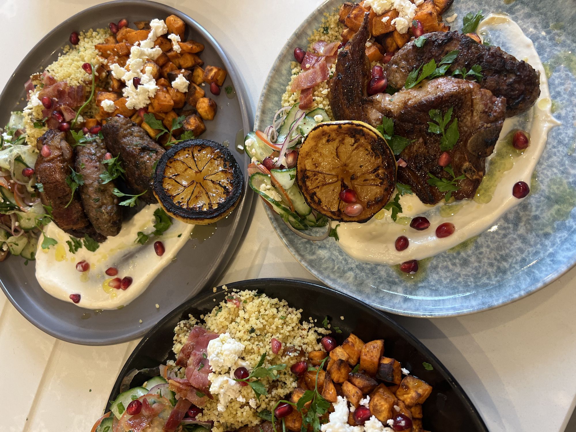 Three plates of food are arranged on a light-colored surface. The plates are filled with a colorful assortment of couscous, roasted sweet potatoes topped with feta cheese, meat (lamb or beef), a charred lemon half, and a cucumber salad. Pomegranate seeds are scattered across all the dishes. The plates are gray, blue, and black. The setting suggests a restaurant environment. Due to the nature of the food style on the plate, it is likely the location is within the city of London, United Kingdom.