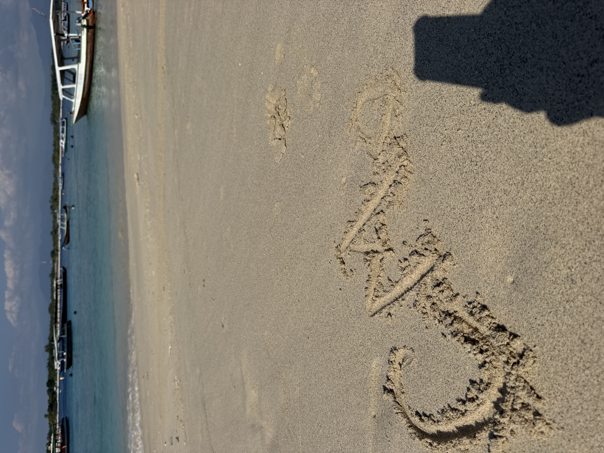 The image depicts a sandy beach with the word "CALA" inscribed in the sand in capital letters. The letters are formed by displacing the wet sand, creating raised edges. In the foreground, the shadow of a person holding a phone is cast on the sand to the right of the inscription.
The background of the image shows a tranquil ocean with several boats anchored or moving slowly. The water is a clear turquoise color, and the sky above is a bright blue with scattered white clouds. The horizon is dotted with a distant shoreline, possibly an island, with some vegetation and mountains visible. The overall scene suggests a peaceful and sunny day at a tropical beach location in Indonesia.