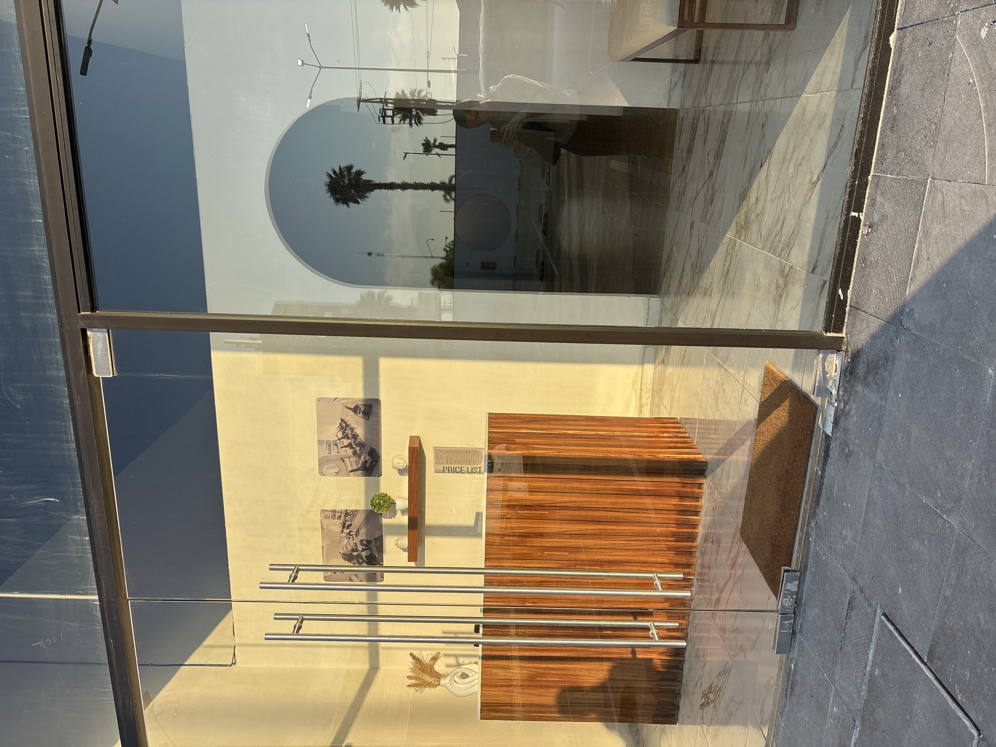 The image depicts the exterior of a storefront in San Bernardino Tlaxcalancingo, Mexico. The entrance is a glass door with multiple horizontal metal handles. Reflected in the glass are palm trees and streetlights, suggesting an urban environment. Inside, a wooden structure with horizontal slats is visible, potentially a counter or shelving unit. The interior wall displays a "PRICE LIST" sign and black and white photographs. A doormat sits just inside the entrance. The exterior pavement is made of dark gray square tiles. A person is reflected in the glass, holding a phone, presumably taking the photograph.
