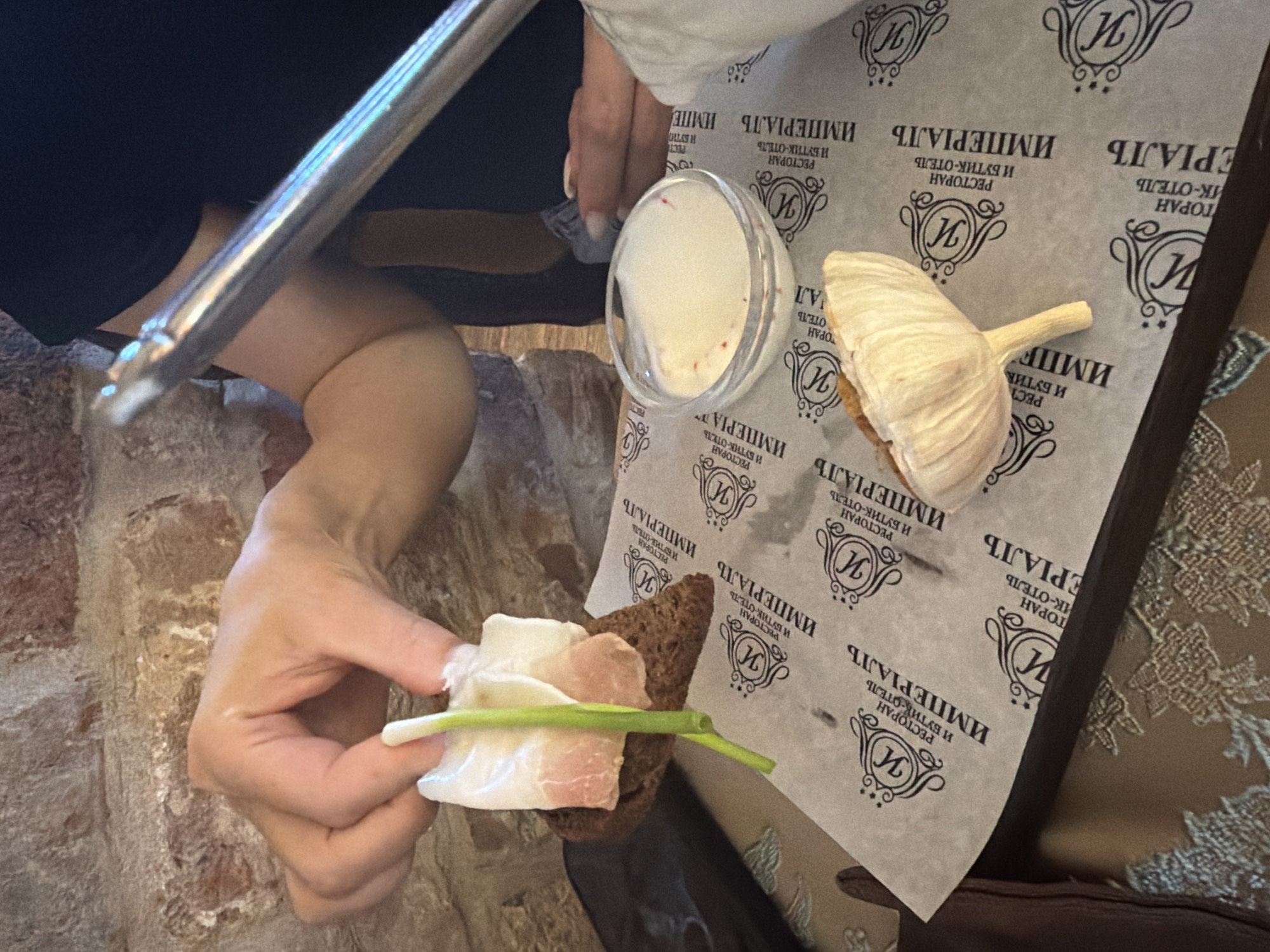 A person's hands are holding a piece of dark bread topped with white fat and a green scallion. The bread is on a piece of paper with a repeating logo. In the background, a small glass bowl contains a white dip with red flecks. To the right, a large clove of garlic is visible. The setting appears to be a restaurant, possibly in Suzdal, Russia, with a brick wall in the background.