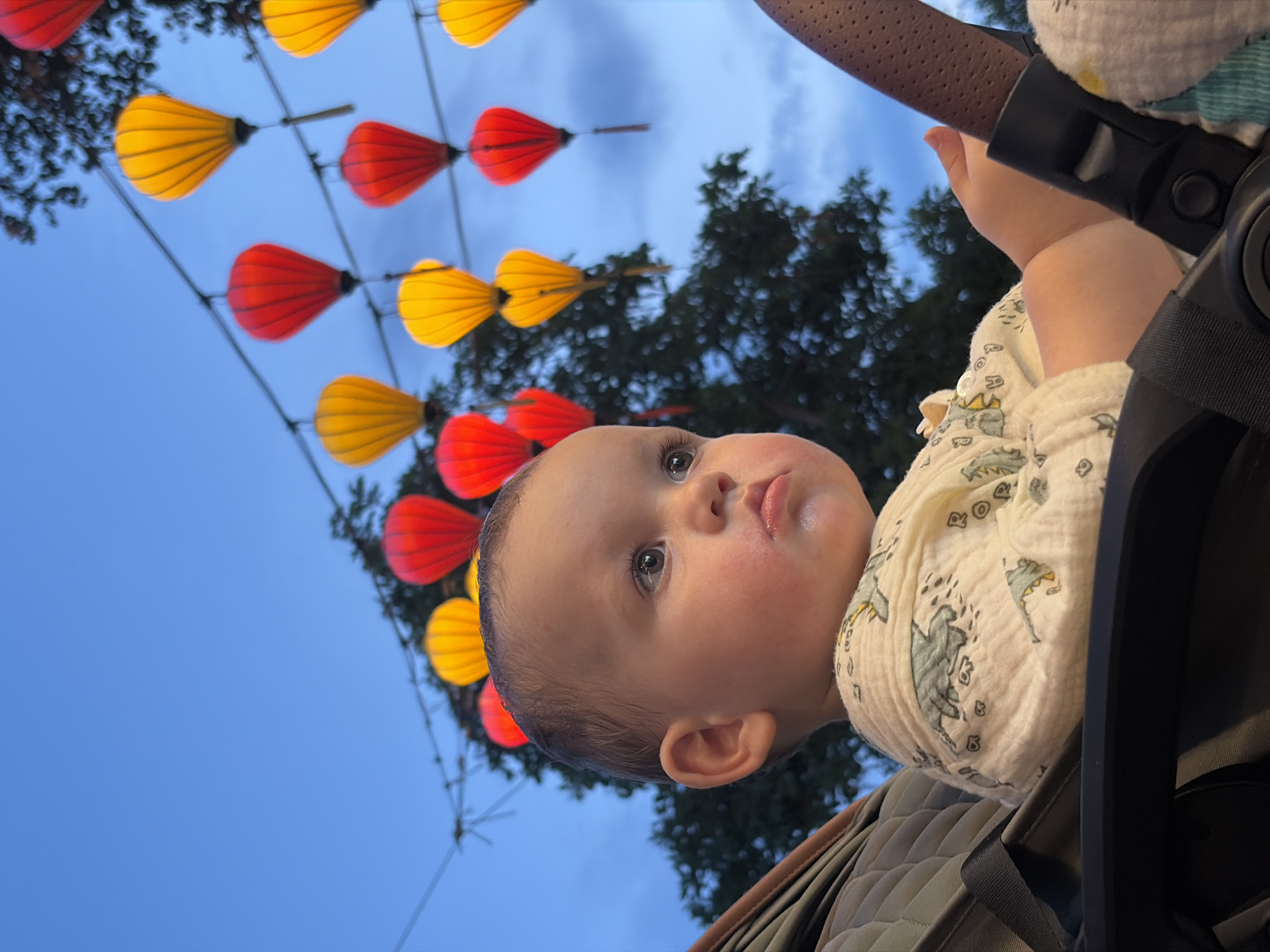 This image captures a close-up, low-angle perspective of a baby looking upwards at an array of decorative lanterns. The baby, situated in a stroller or carrier, occupies the lower right portion of the frame, with their head slightly tilted and their eyes wide open, gazing towards the left side of the image. They have fair skin, dark eyes, and a neutral, curious expression. A light-colored, possibly muslin, patterned shirt with subtle grey designs is visible on their torso.
Above the baby, a canopy of vibrant red and yellow lanterns hangs across wires, stretching from the upper left to the middle of the frame. These lanterns are shaped like elongated teardrops or petals and are illuminated, casting a warm glow. The sky, a prominent feature, is a deep blue, indicative of dusk or early evening, with soft, sparse clouds. Dark silhouettes of trees are visible beneath the lantern strings and against the sky, suggesting an outdoor setting.
Parts of the baby's stroller or carrier are visible on the right and bottom right, including a brown, textured handle, a black frame element, and quilted fabric. The distinctive style and arrangement of the lanterns strongly suggest the cultural ambiance of Hội An Ward, Vietnam, where such displays are common. The overall scene conveys a serene and enchanting atmosphere as the baby observes the illuminated decorations. There is no visible text in the image.