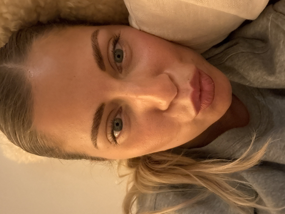 A fair-skinned woman with blonde hair is resting against a white pillow and wearing a gray hoodie. Her eyes are light green. Her makeup is natural, enhancing her eyebrows and lips. The lighting is soft, casting subtle shadows across her face. The overall impression is one of calmness and repose.