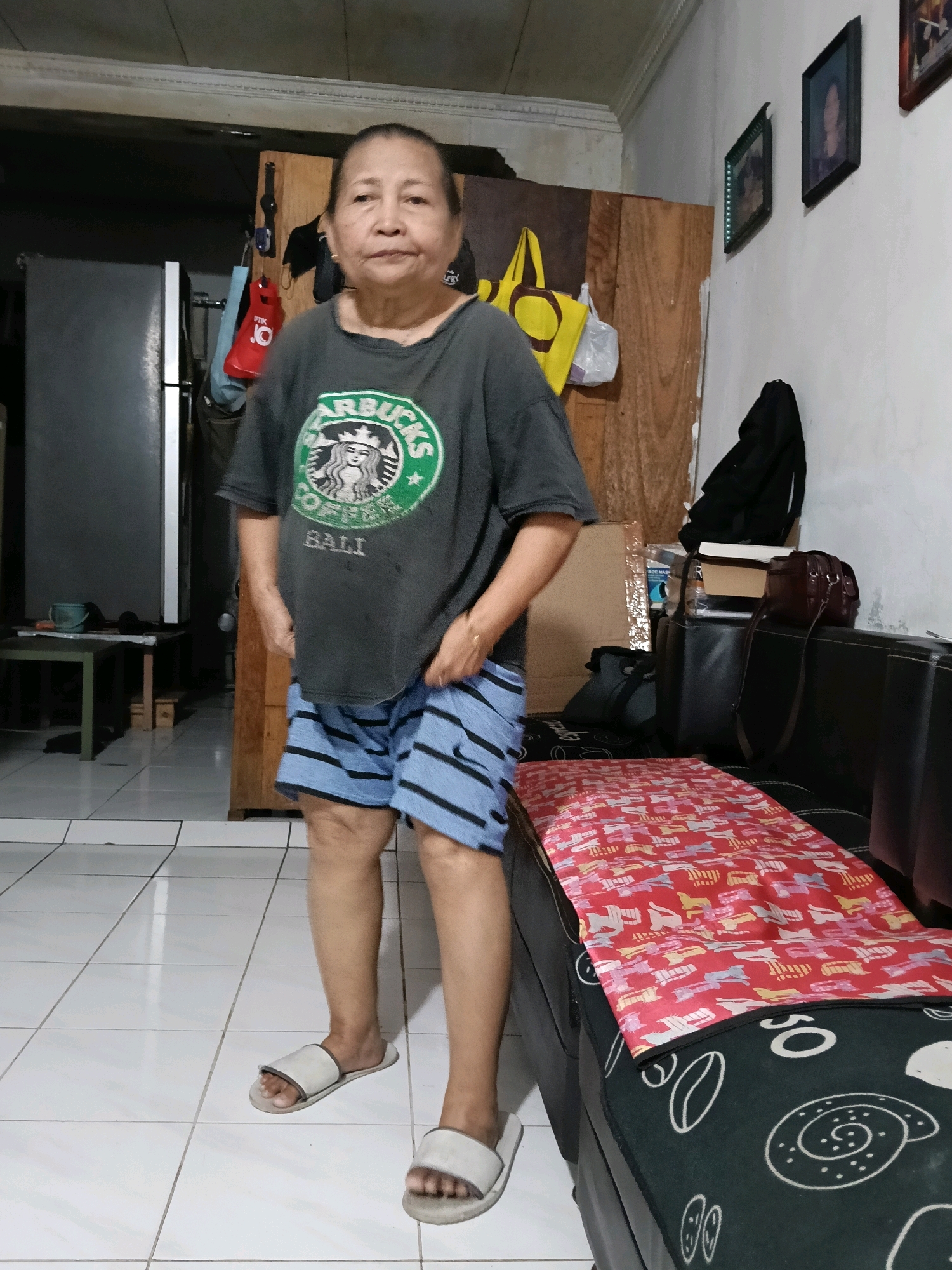 An Indonesian woman stands in what appears to be a living room or common area. She is wearing a dark grey t-shirt with a Starbucks logo and "BALI" printed on it, along with blue and white striped shorts and light-colored slip-on sandals. The woman is positioned in the center of the frame. In the background, a refrigerator is visible to the left, and a wooden cabinet with hanging items such as bags and clothing is to the right. The room has tiled flooring and white walls adorned with framed pictures. A black sofa with a patterned throw is on the right side of the image. The scene suggests a casual domestic setting within Jatimulya, Indonesia.