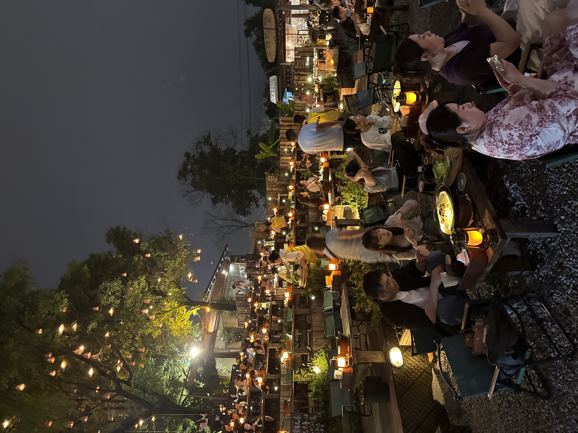 The image depicts an outdoor dining area in the Nan'an District of China, during nighttime. Numerous people are seated at tables, some using their cell phones. Each table is illuminated by a lantern. The area is decorated with trees wrapped in strings of small lights. Some of the seating includes folding chairs. The ground around the tables appears to be covered in gravel. Buildings are visible in the background.