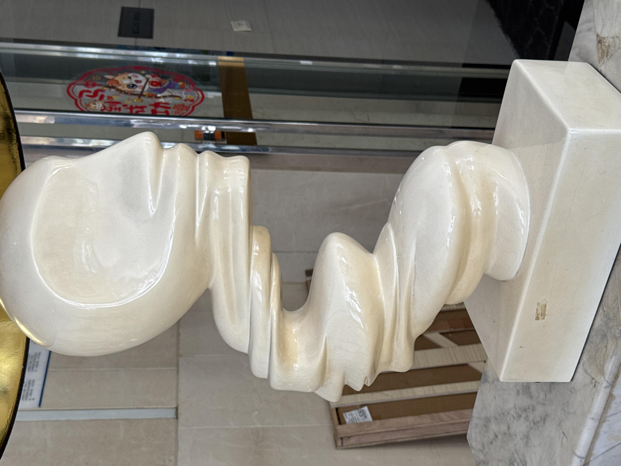 A porcelain sculpture is featured prominently in the image. The sculpture appears to be a white color, with an elongated, twisted shape. It's placed on a marble-like surface. Above the sculpture, there's a metal rack or shelf, and on that shelf, a sticker with Chinese writing is visible. The location is identified as Yuzhong District, China, based on image metadata or surrounding information. No people or actions are visible. The focus is solely on the sculpture and its immediate surroundings.