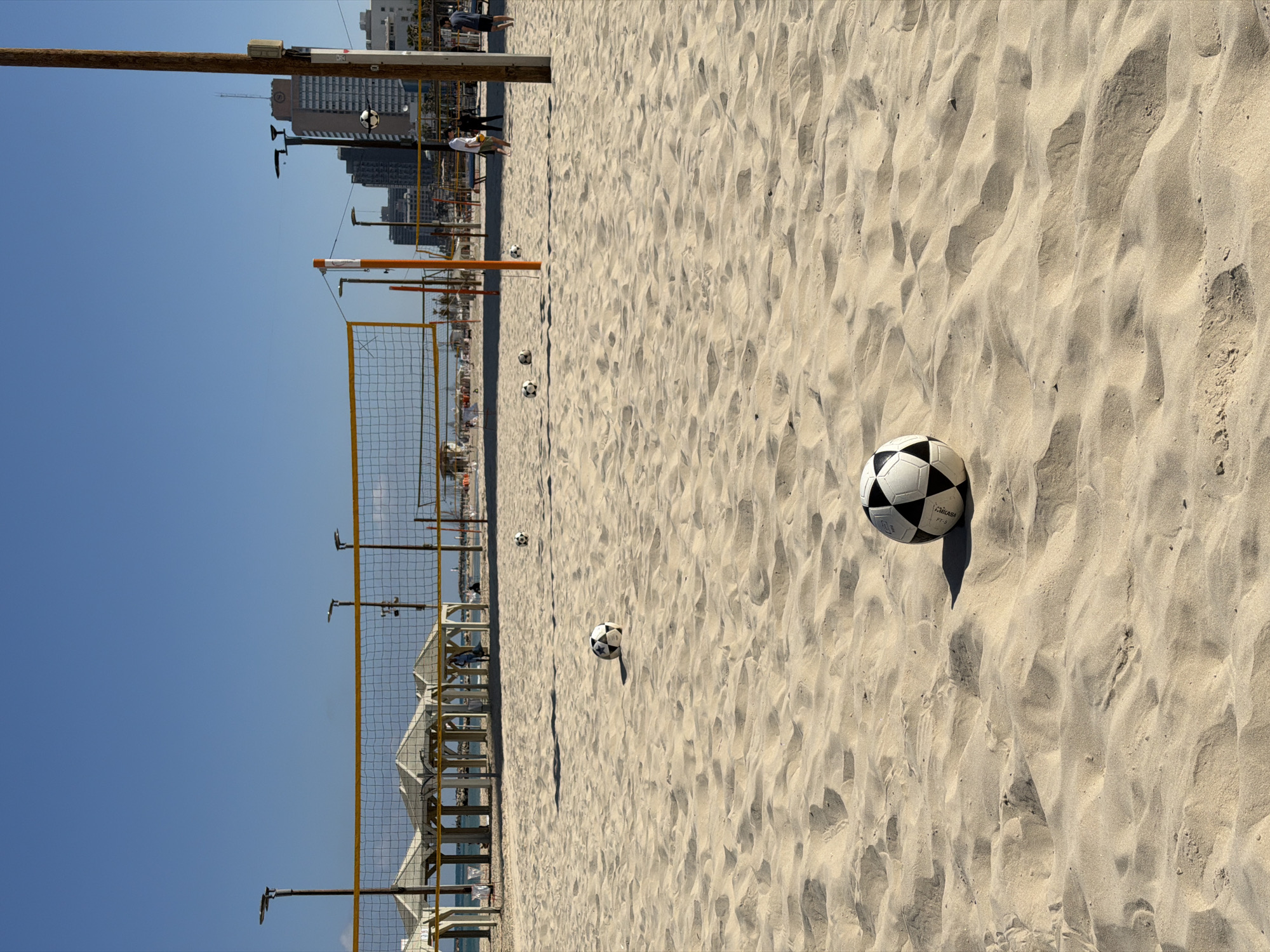 The image depicts a beach in Tel Aviv, Israel, featuring a volleyball net and multiple soccer balls scattered across the sand. In the background, there are several buildings, utility poles, and people walking on the beach. The sky is clear and blue. The sand displays a pattern of shadows and highlights.