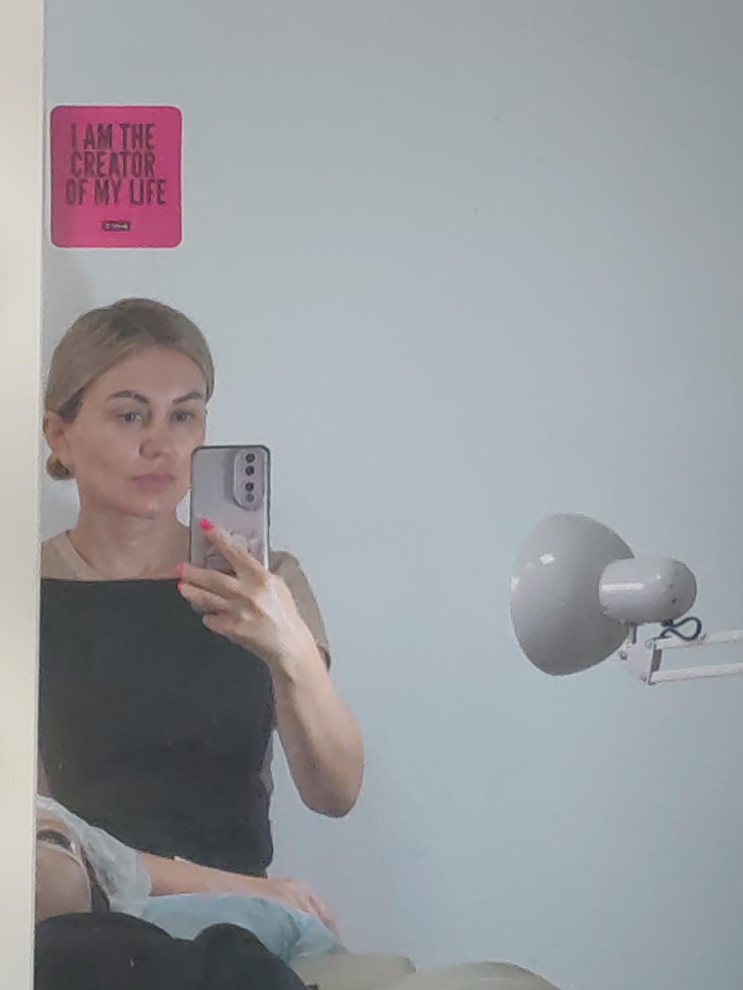 The image shows a woman taking a mirror selfie in what appears to be a domestic or personal care setting. She is holding a phone with a pink case and has bright pink fingernails. She is wearing a tan t-shirt and a black top over it. Behind her, on the wall, is a pink square sticker with black text that reads "I AM THE CREATOR OF MY LIFE". To the right of the woman, a white desk lamp with a round shade is visible. The background is a plain, light-colored wall. The image composition suggests the photograph was taken in Komsomolsk-on-Amur, Russia, based on the prompt's instruction.