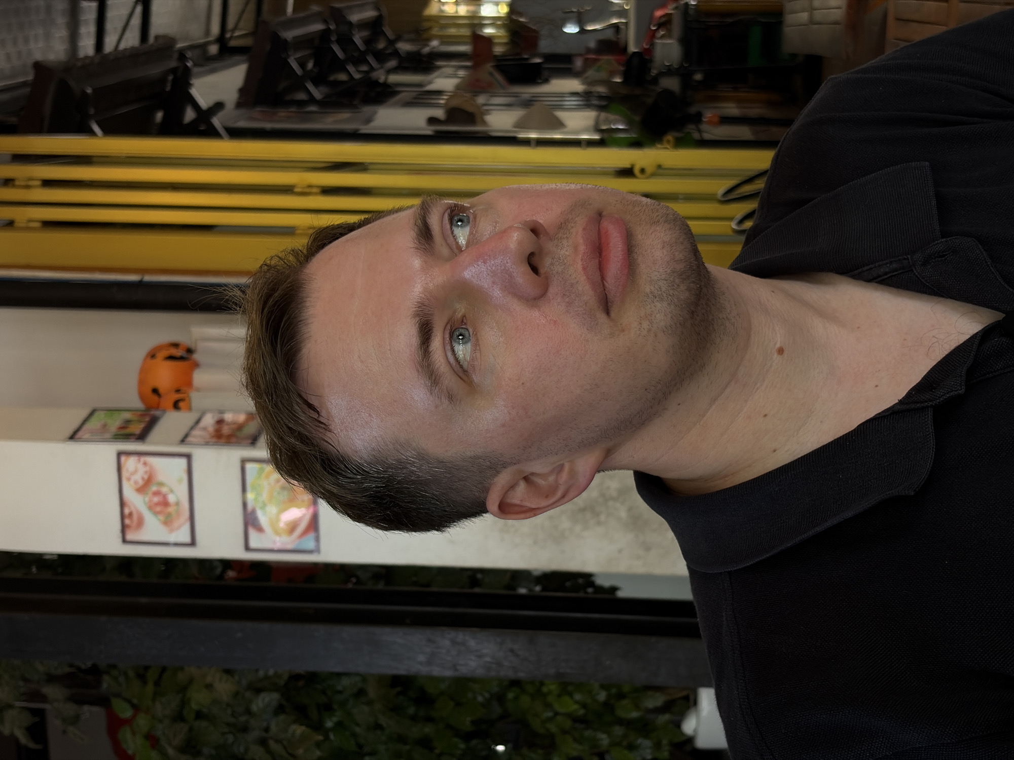 The image presents a close-up portrait of a man, rotated 90 degrees clockwise. He has a fair complexion, distinct blue-green eyes, short-cropped brown hair, and light stubble along his jawline and chin. He is wearing a black collared polo shirt. His gaze is directed calmly upwards and to the viewer's right, with a neutral and composed expression.
The background suggests an indoor or semi-covered setting, likely a cafe or restaurant in Đà Nẵng, Vietnam. Behind the man, a series of prominent horizontal yellow metal beams or decorative slats are visible. To the far left, a small, bright orange object resembling a decorative pumpkin or Halloween ornament is present. On a white wall behind the man's head, several framed illustrations or photographs depict various food items, including what appear to be tacos or salads, further reinforcing the dining establishment context. A dark, heavy beam or ledge crosses the lower portion of the frame, with some green foliage, possibly artificial, visible beneath it. The scene is evenly lit, indicating indoor illumination. No specific text is clearly legible on the background elements.