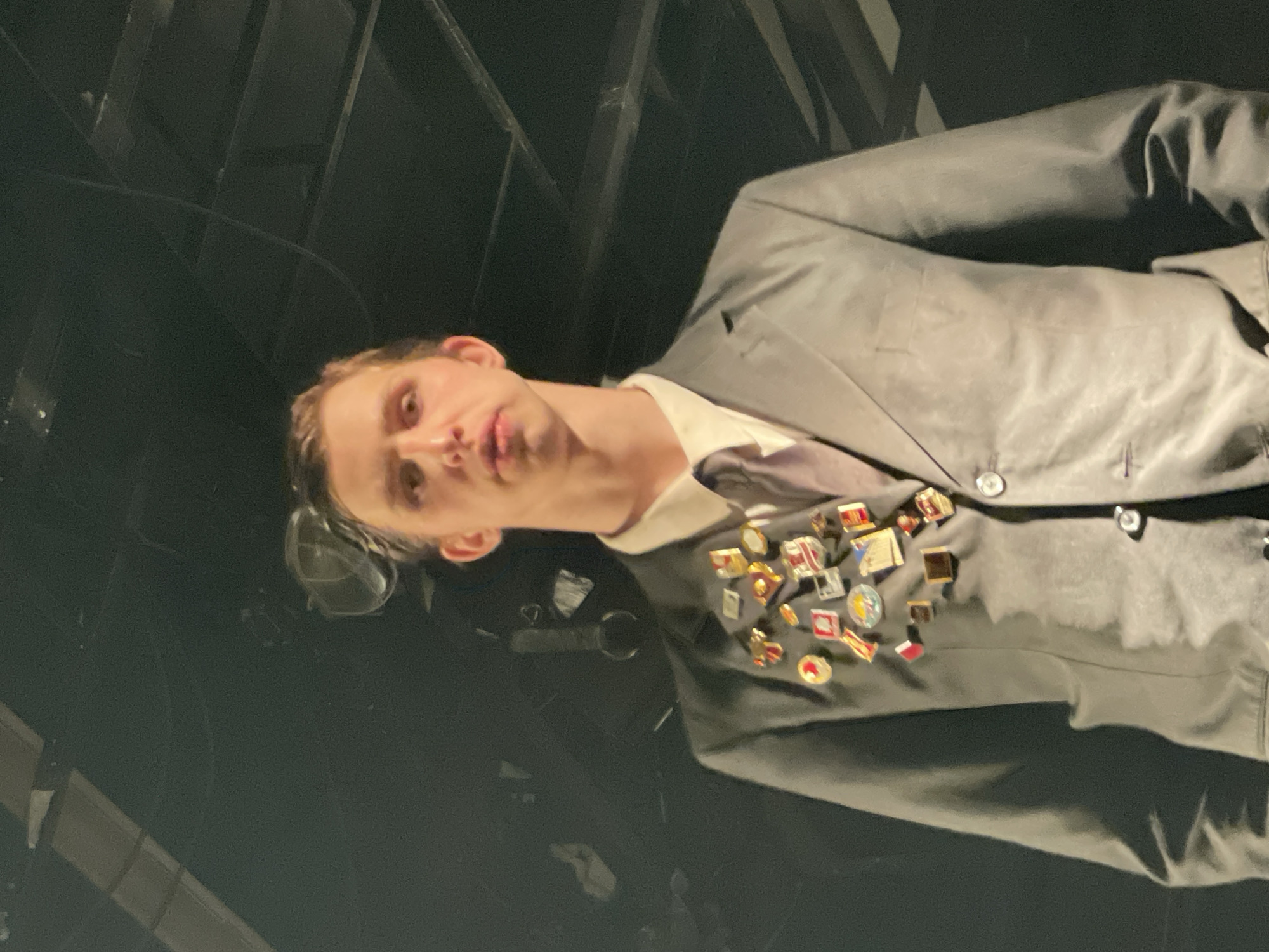 A man in a gray suit, white shirt, and gray tie stands in front of a dark background that shows the edges of what appear to be metallic bars or a fence. The man's suit jacket is covered in numerous small pins and badges on the lapel area. The setting appears to be indoors, although the location is ambiguous. Moscow, Russia is not discernible from the image.