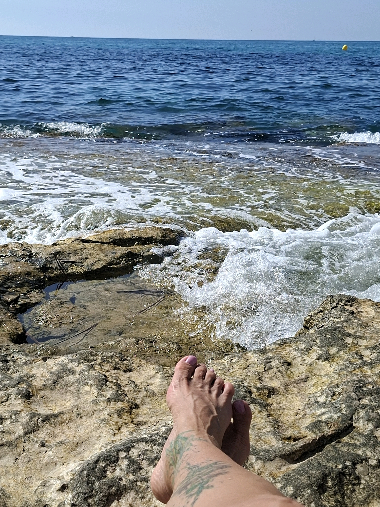 The image captures a serene coastal scene with a person's feet resting on rocky terrain overlooking the Mediterranean Sea. The foreground is dominated by the textured, sun-bleached rocks, some featuring shallow pools of water reflecting the surroundings. A wave is breaking in the middle ground, creating white foam and spray as it washes over the rocks. The sea stretches out to the horizon, its blue waters gently rippling under a clear sky. A yellow buoy is visible in the distance. The person's feet, adorned with light green tattoos on the left leg, are positioned towards the bottom of the frame. The overall atmosphere is one of tranquility and relaxation by the sea, consistent with a location like Torrevieja, known for its coastline.
