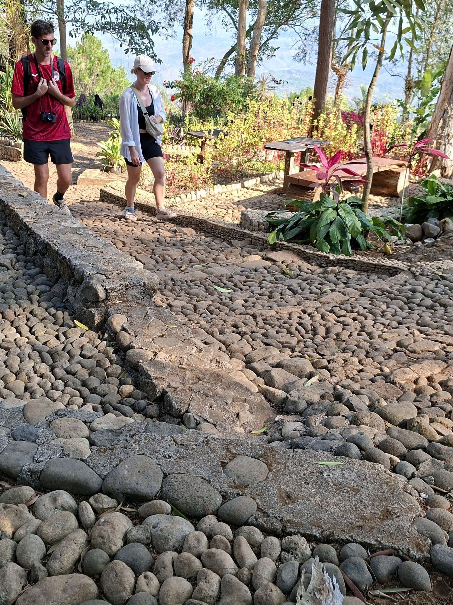 In the city of Dlingo, Indonesia, a man and a woman are walking on a stone path surrounded by lush greenery and trees. The man, wearing a red t-shirt and black shorts, has a camera around his neck. The woman, in a white shirt over a black top and shorts, wears a white cap and a beige fanny pack. They are walking downhill on a path made of rounded river stones, bordered by a low stone wall. In the background, a wooden table and benches are visible, set amidst vibrant flowering plants and foliage, with a backdrop of distant hills and trees. The lighting suggests it is daytime, with shadows indicating a sunny environment.
