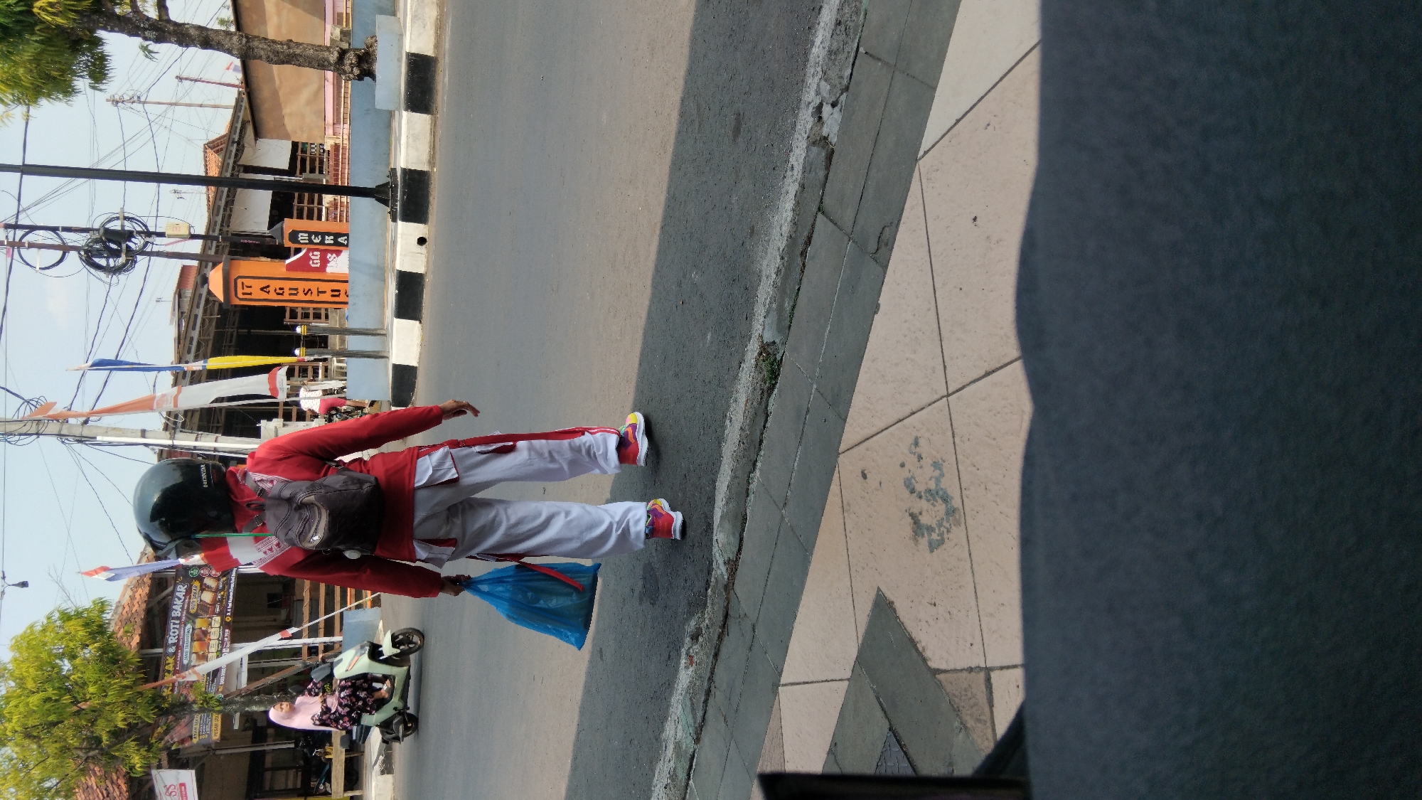 A person wearing a red jacket, white pants with red stripes, a black helmet, and a backpack is walking on a street in Batang, Indonesia. They are carrying a bright blue plastic bag in their left hand. The person is walking away from the camera, towards a building with signage that appears to be a food establishment, indicated by the "ROTI BAKAR" (grilled bread) text. In the background, there are flags, a traffic light pole, and other pedestrians. To the left of the main subject, a woman on a light green scooter is also visible. The environment suggests a sunny day with shadows cast on the sidewalk and street. The scene is set on a paved road with a sidewalk made of checkered tiles. A portion of a dark, textured surface, possibly a table or bench, occupies the foreground.