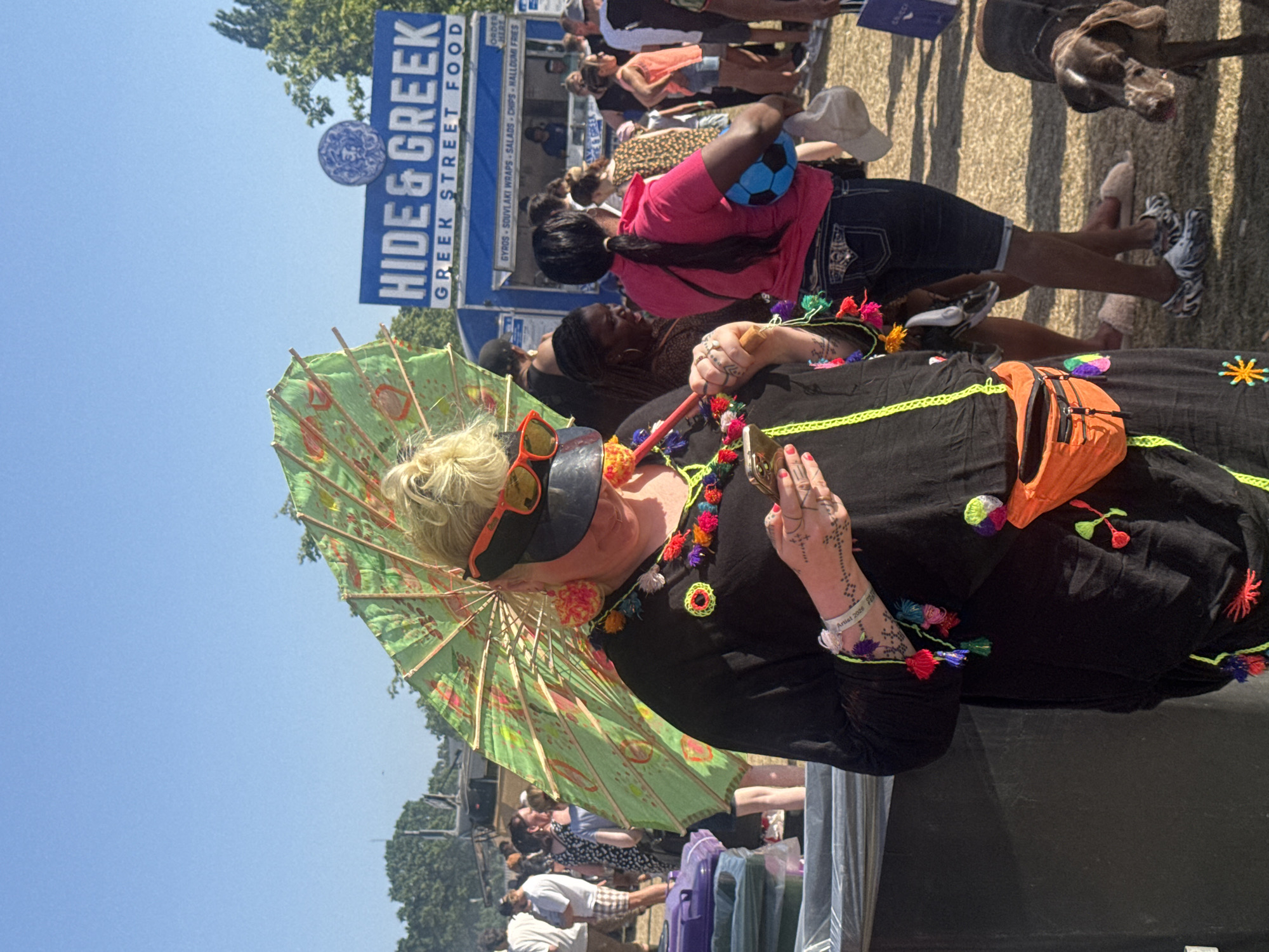 The image depicts a woman holding a parasol in a crowded outdoor environment. She is wearing a black dress embellished with colorful tassels and an orange fanny pack. Her accessories include orange sunglasses and wrist tattoos. In the background, a food vendor stall called "HIDE & GREEK" indicates that the location is a street food event, with a sign advertising Greek street food such as gyros, souvlaki wraps, salads, chips, and halloumi fries. Other people can be seen in the background, some waiting in line at the food stall. There is also a dog visible. The weather appears sunny and clear, judging by the bright blue sky. Based on the "Artist 2026" wristband, it is likely a music or arts festival. Since the OCR text includes the phrase "HALLOUMI FRIES" and the location is the city of London, United Kingdom, I deduce it is the city of London, United Kingdom, and there is halloumi fries for sale.