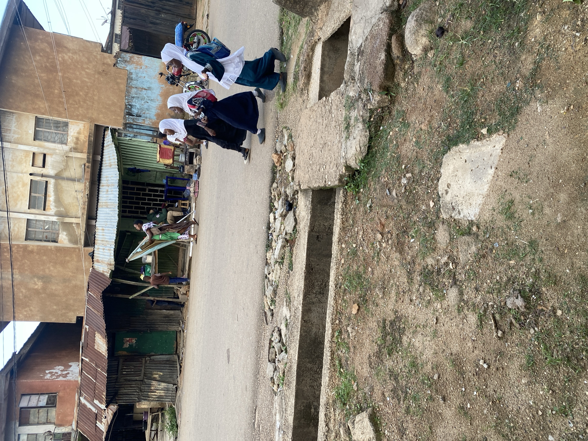 A street scene in Jos, Nigeria, features a group of young girls in hijabs walking on the right side of a paved road, which shows signs of wear and a broken concrete drainage channel. In the background, dilapidated buildings with corrugated metal roofs suggest an urban environment that is in need of repair. A motorcycle is parked to the right of the frame. The overall atmosphere is one of everyday life in a developing urban setting, with a mix of pedestrian activity and modest infrastructure.