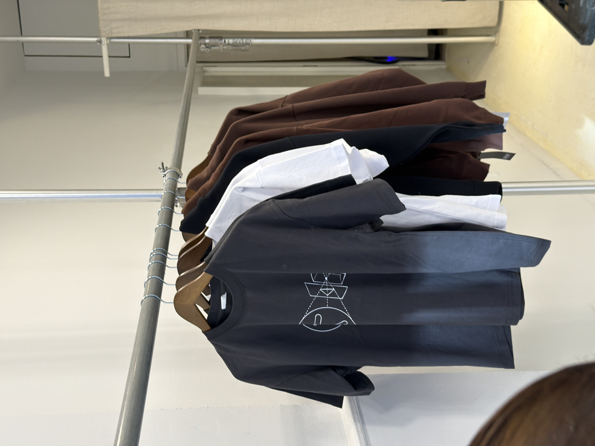 A rack of clothing is displayed in what appears to be a retail setting. On the rack, there are several items, including brown hoodies, black clothing items, white t-shirts, and dark gray or black t-shirts featuring a graphic design. The graphic design on the dark t-shirts includes geometric shapes and an eye-like symbol. The clothing is suspended from wooden hangers. The rack itself is constructed of metal and has a simple, industrial design. The background features a neutral-colored wall and ceiling, as well as a partial view of a light-colored curtain. The lighting is bright, and the scene seems well-lit. The location of the photo is not evident from the image alone.