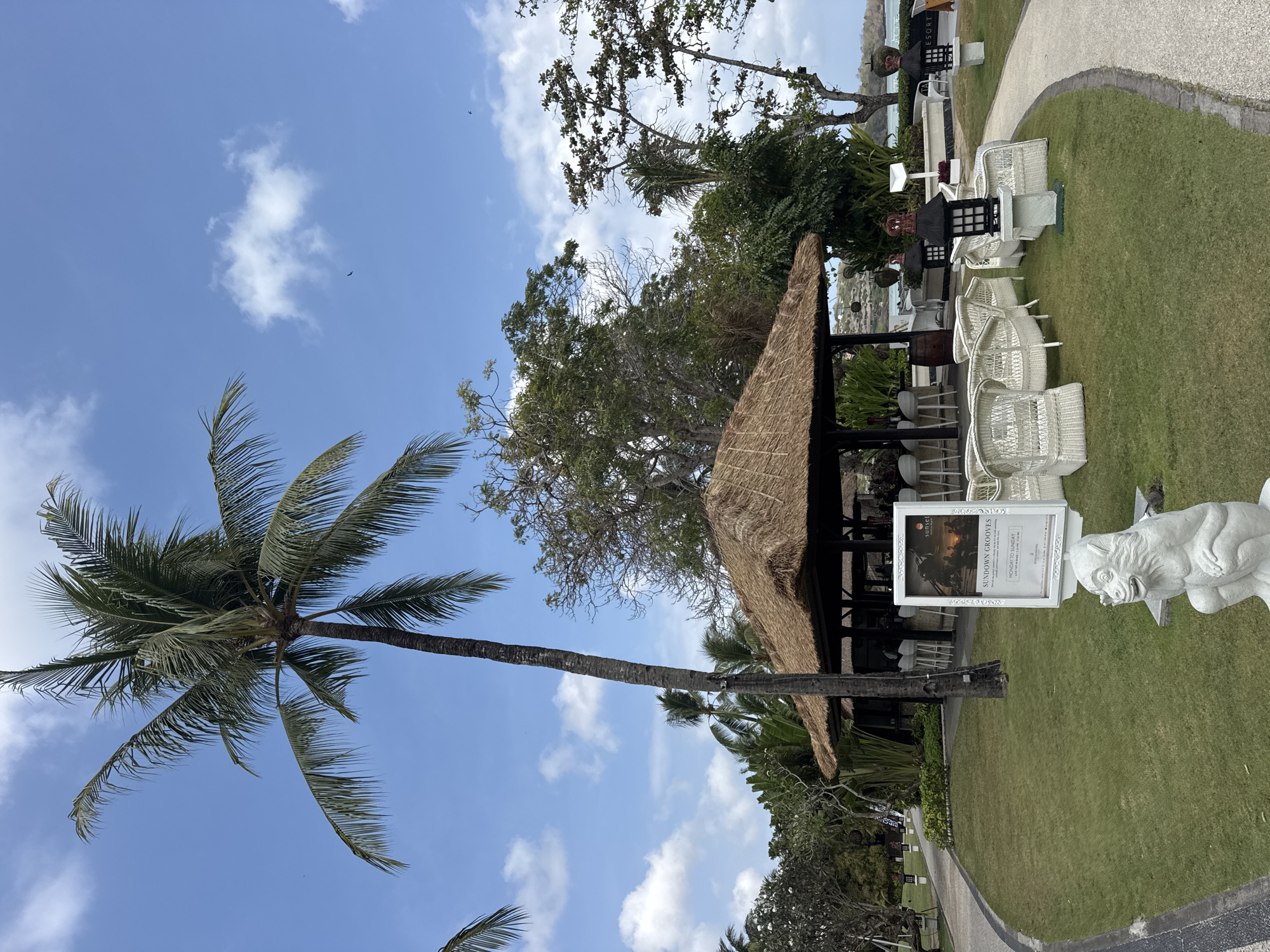 A resort bar area is pictured on a sunny day in Kuta Selatan, Indonesia. The scene is dominated by a thatched-roof pavilion housing a bar with white stools. In front of the bar, white wicker chairs are arranged on a manicured lawn. A path curves around the lawn, leading past a sign advertising "Sundown Grooves" and a white stone monkey statue. A tall palm tree stands to the left, and lush tropical foliage fills the background. The sky is blue with scattered white clouds.