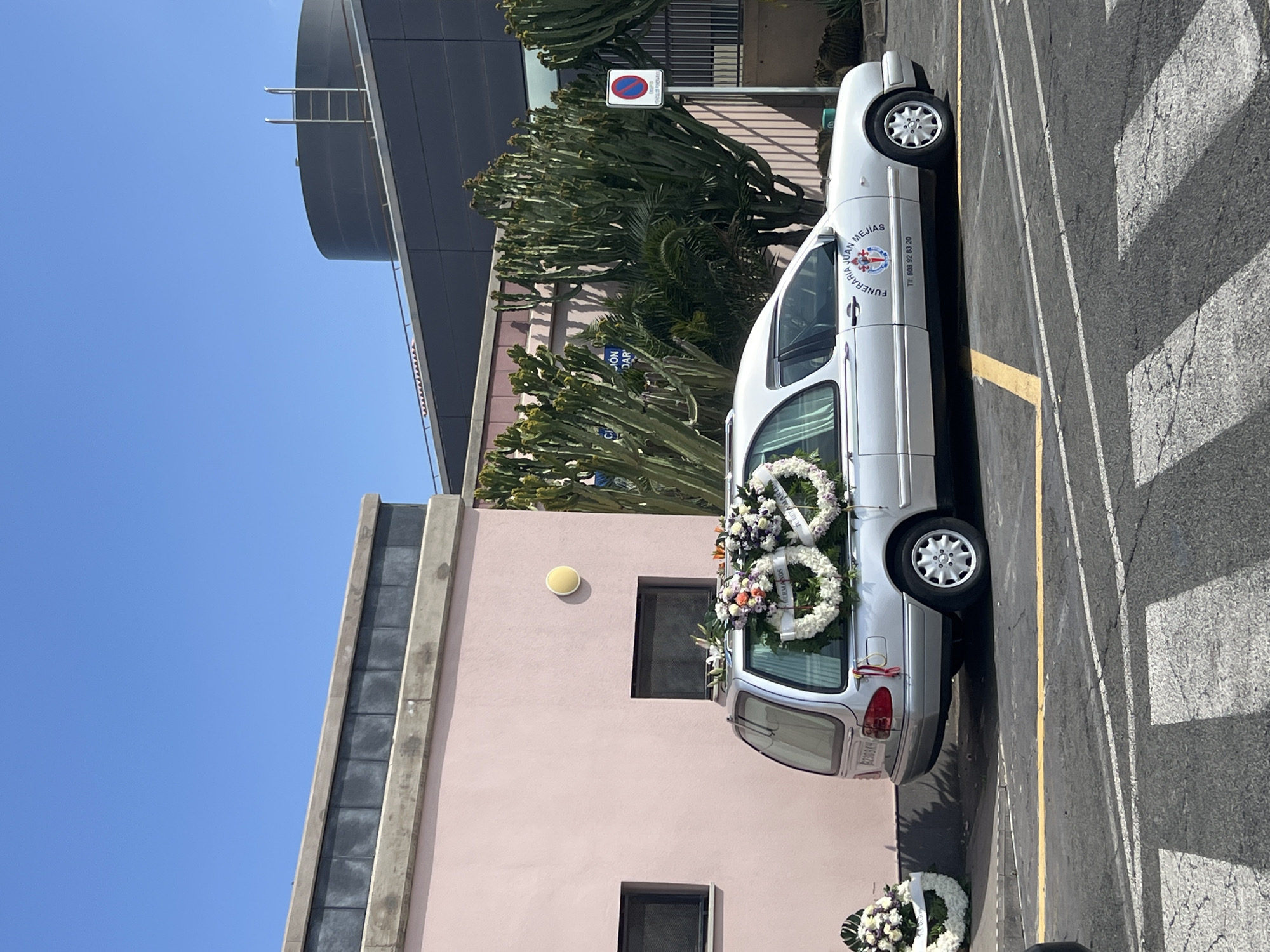 A silver Mercedes-Benz hearse, operated by "FUNERARIA JUAN MEJIAS" with the contact number Tlf: 608 92 83 20 displayed on its side, is parked facing right in a paved lot in Cruce De Sardina, Spain. The vehicle's rear cargo area is laden with numerous floral wreaths, predominantly white and purple, including two distinct heart-shaped white wreaths, one inscribed "HERMANDAD" and the other "H. LUNA MANUEL." An additional white floral arrangement rests on the ground adjacent to the hearse's left rear. The scene unfolds under a clear blue sky.
To the left, a light peach-colored building with two dark rectangular windows and a yellow circular wall light is visible. Behind the hearse and extending to the right is a multi-story building featuring a darker facade, partially obscured by several tall, multi-stemmed euphorbia plants. A 'No Parking' sign, bearing the additional text "EXCEPTO VEHICULOS FUNERARIOS" (Except Funeral Vehicles), stands near this darker building. A cylindrical structure with a metal ladder is observed on the roofline of the darker building. The paved lot exhibits yellow parking space markings and white pedestrian crossing stripes. No individuals are present in the image.