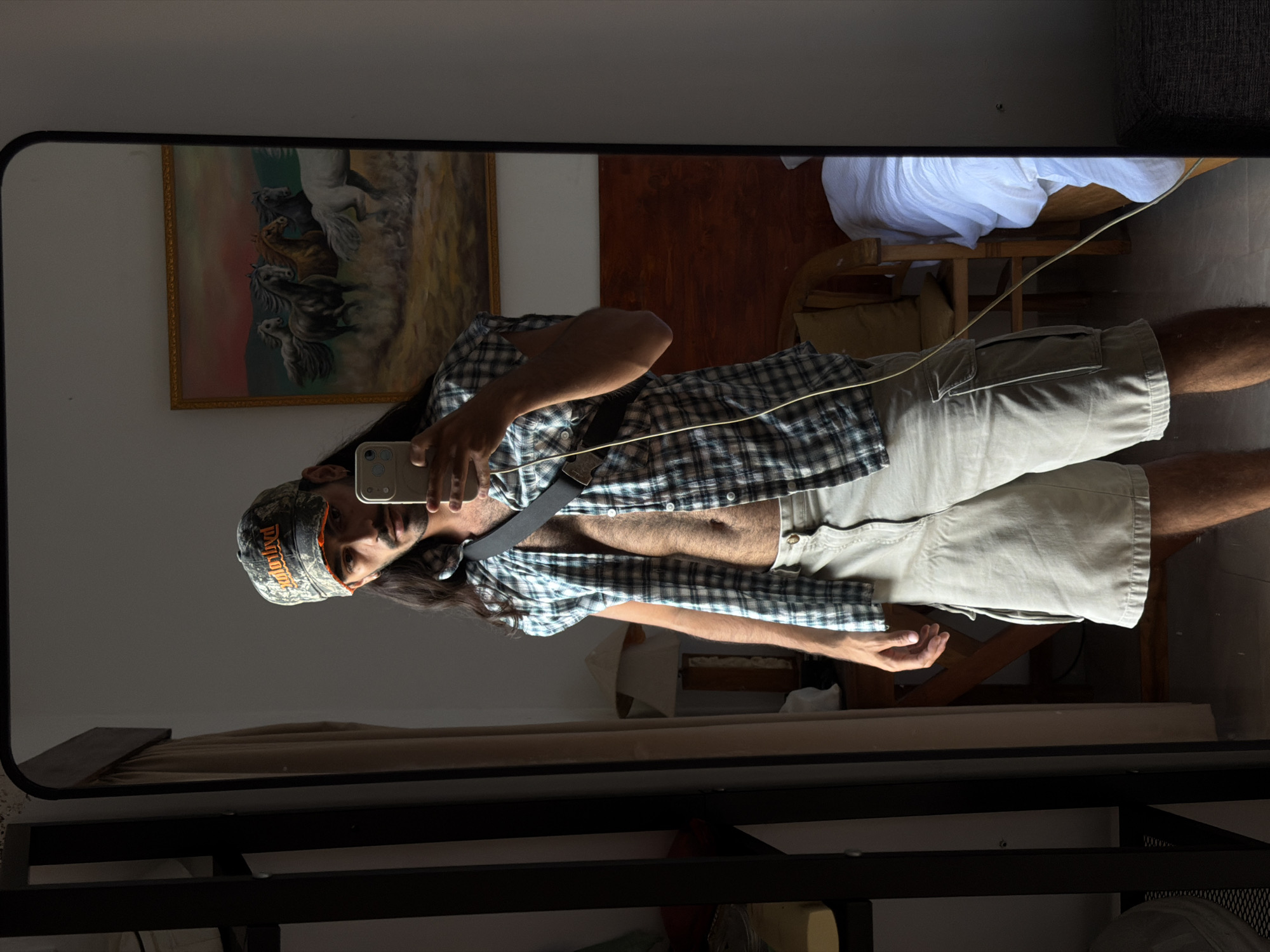 A man is captured in a mirror selfie, standing indoors in what appears to be a room. The image is presented rotated 90 degrees clockwise from its original orientation, so the man is seen horizontally. He has long dark hair and a beard, wearing a light-colored plaid button-up shirt unbuttoned to reveal his chest, light khaki cargo shorts, and a black belt. On his head, he wears a camouflage baseball cap with orange lettering that reads "Harley-Davidson." He holds a light-colored smartphone up to take the photo, with multiple camera lenses visible on its back. A thin, light-colored cable crosses his body.
The reflection in the mirror reveals elements of the room. On the wall to the left, there is a framed painting depicting several horses (white, brown, and dark grey) galloping across a landscape with warm, earthy tones and some green. To the right of the man, a wooden chair is visible, draped with a white cloth. Below the chair, another piece of furniture or pillow is partially seen. The overall lighting suggests daytime, with some areas of shadow, particularly in the upper left of the mirror's reflection. The mirror itself has a thin black frame. Below the mirror's edge, a dark shelf or table structure is visible, holding some objects, including a white mesh item. The setting is an interior space, consistent with a home in North Kuta, Indonesia.