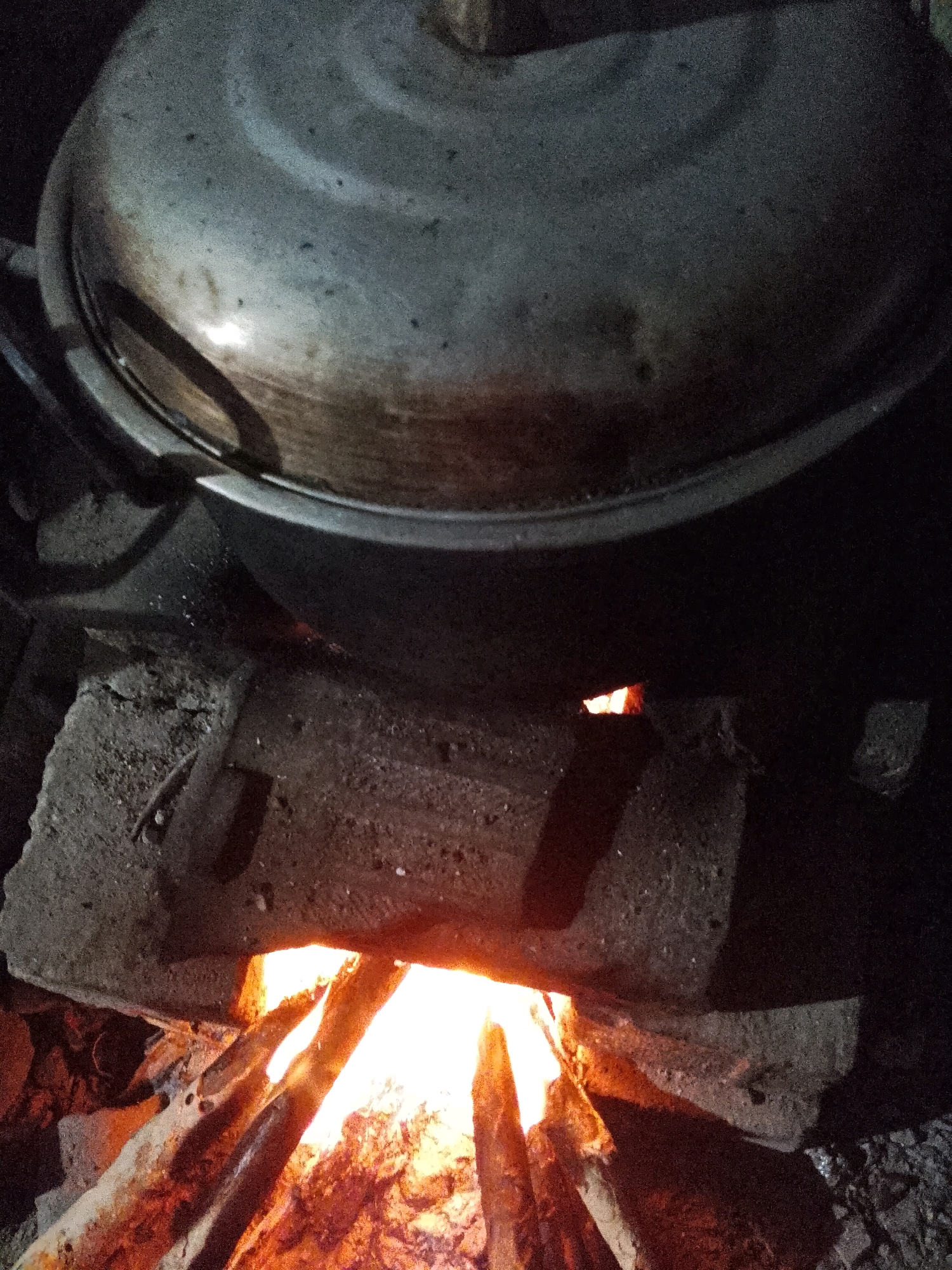 An overhead, medium shot captures a pot covered by a lid, resting on a makeshift stove in Wadang, Indonesia. The stove is constructed from bricks and rocks, with burning wood visible beneath the pot, casting a warm glow on the surroundings. The pot, made of metal, appears well-used, with its surface exhibiting signs of age and perhaps some soot. The scene suggests a domestic setting, possibly for cooking. The lighting is dim, indicating it may be nighttime or an indoor cooking area with minimal illumination. There are no people visible in the frame.