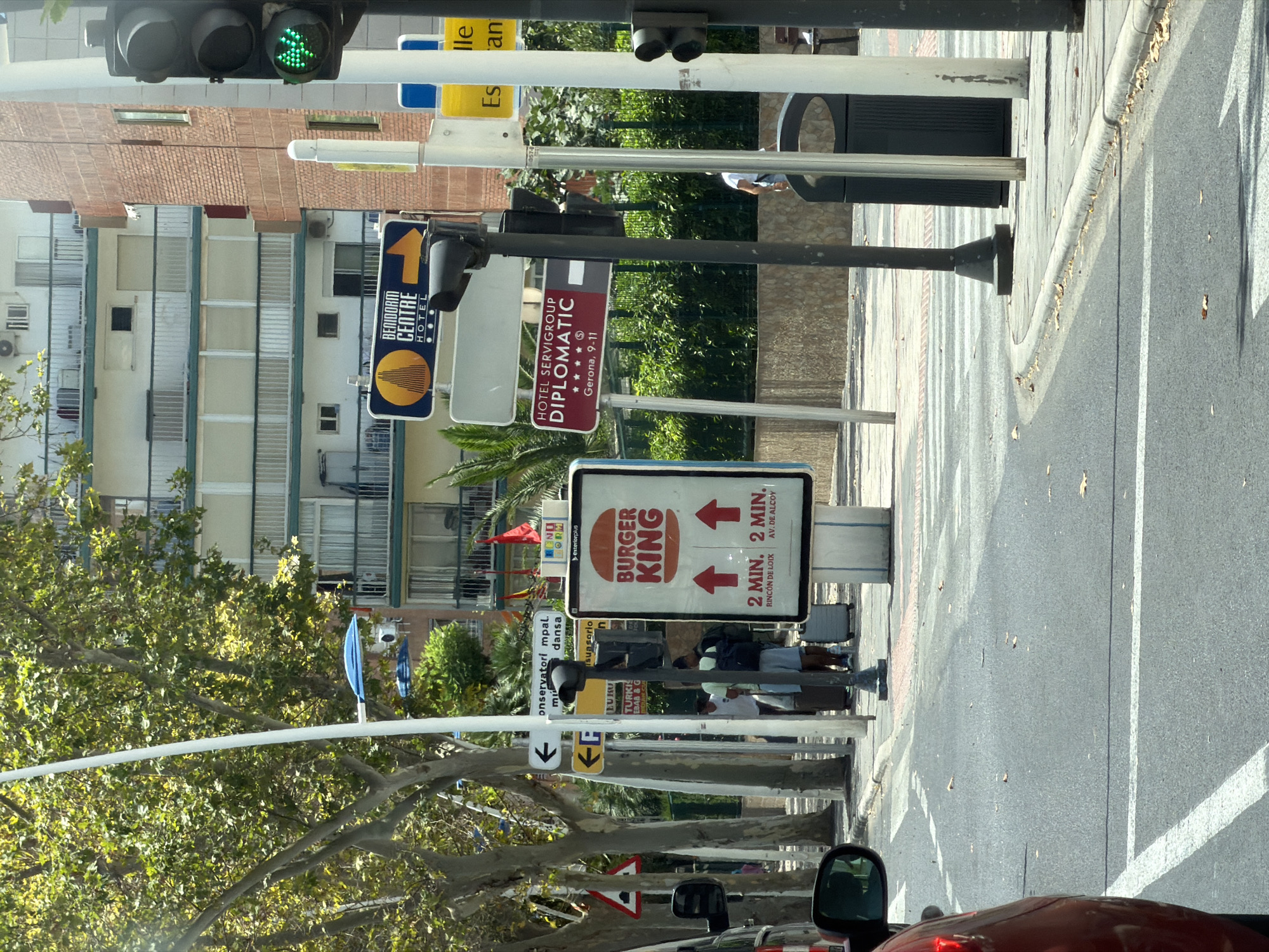 The image captures a street scene in Benidorm, Spain, on a sunny day. A Burger King advertisement sign is prominently displayed, showing the restaurant's logo and advertising a 2-minute wait time for food with arrows pointing upwards, indicating direction. Nearby, several directional signs are visible, including one for "BENIDORM CENTRE HOTEL" with a yellow arrow pointing to the right, and another for "HOTEL SERVIGROUP DIPLOMATIC" with an address in Gerona. A traffic light with a green signal is also present. In the background, a multi-story building with balconies and apartments lines the street. Trees with lush green foliage provide shade along the sidewalk. Several individuals are seen in the scene, some with luggage, suggesting they might be tourists. A red car is partially visible in the foreground, indicating that the photo was taken from within a vehicle. The street is paved, and a pedestrian crossing is marked on the road. A black trash bin is located on the sidewalk.