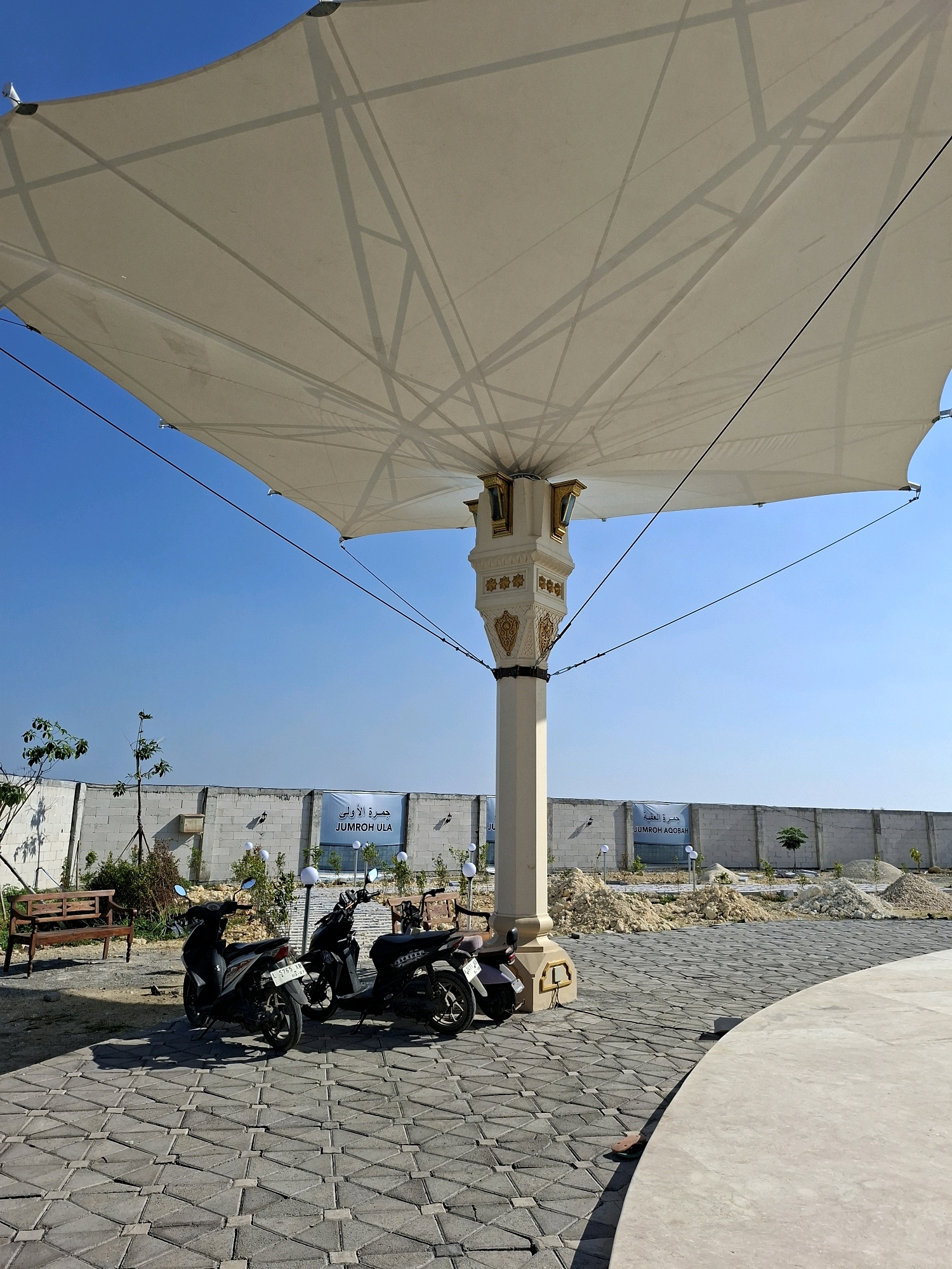 In Jrengik, Indonesia, under a large, white tensile fabric umbrella supported by a decorative pillar, two parked scooters are visible on a paved area. The ground is covered with interlocking grey concrete pavers arranged in a geometric pattern. In the background, a white brick wall stretches across the scene. Behind the wall, piles of sand and gravel indicate ongoing construction. Two banners are affixed to the wall; one displays Arabic text and "JUMROH ULA," while another shows Arabic text and "JUMROH AQOOBAB." A wooden bench sits to the left of the scooters, and sparse vegetation, including small trees and bushes, is present. The sky above is clear and blue.