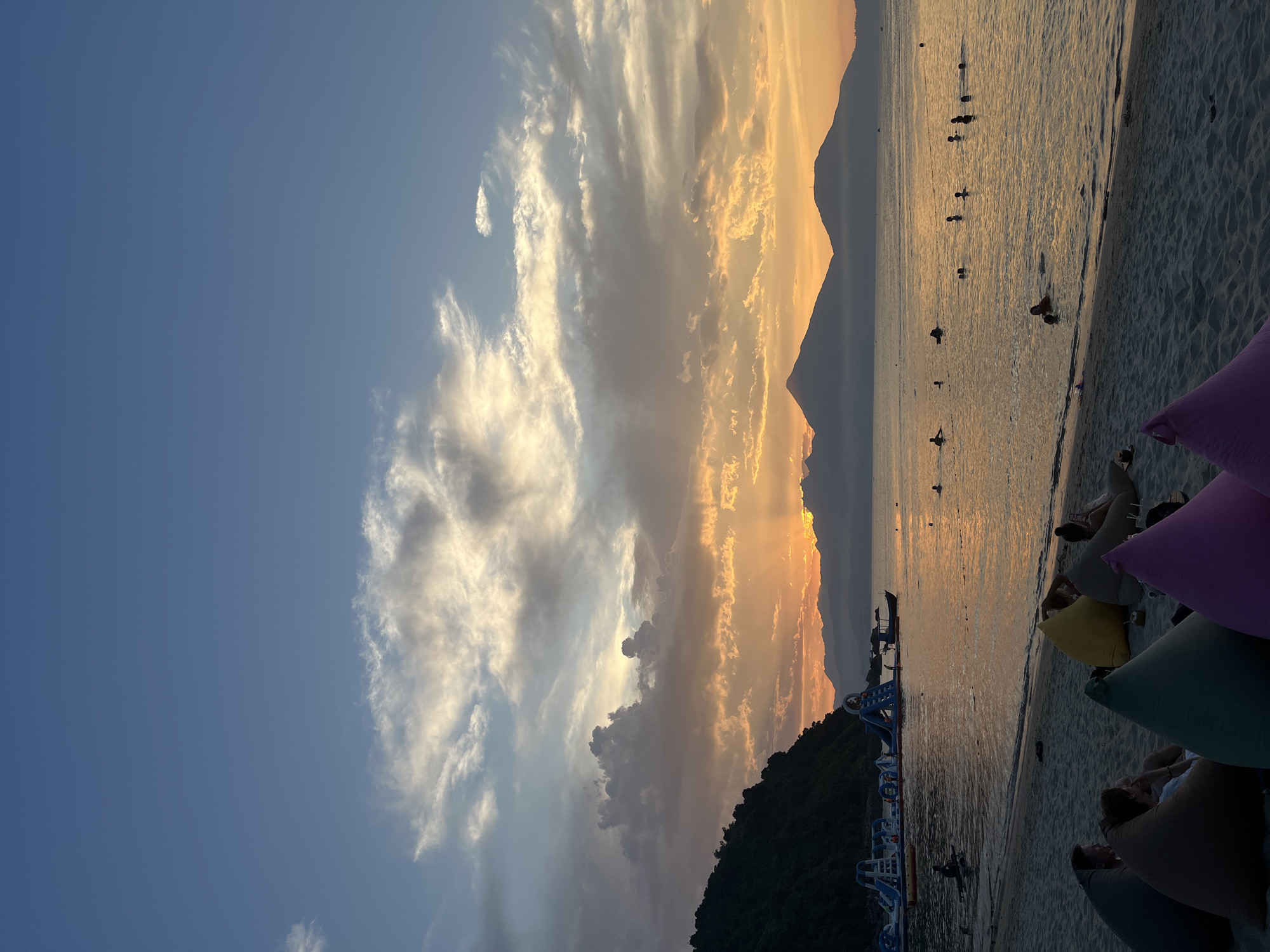The image depicts a beach scene at sunset, possibly in Đà Nẵng, Vietnam, given the presence of distant mountains and coastal structures. The sky is a mix of blue and golden hues, with clouds illuminated by the setting sun. The beach has a sandy shoreline with several people in the water and on the sand, some appearing to be swimming or wading. There are floating water park structures visible along the coastline. In the foreground, there are several beanbag chairs, some of which are occupied by people relaxing on the beach. The scene suggests a calm, late-day atmosphere at the beach.