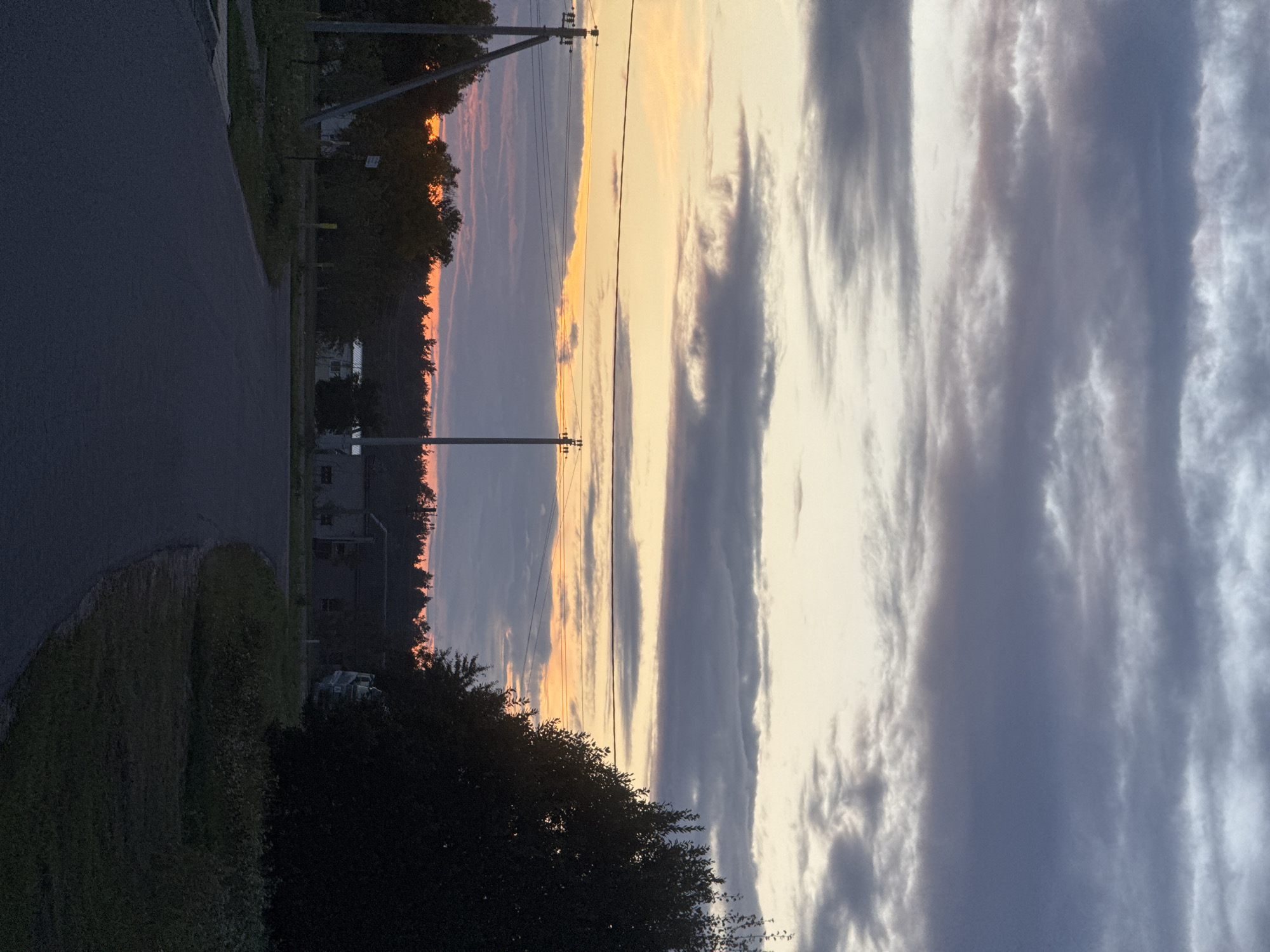 A rural road curves through a landscape under a twilight sky. The sky is dominated by layered clouds, with streaks of orange and pink near the horizon, indicating sunset or sunrise. Power lines stretch across the upper portion of the image, supported by utility poles visible on the left and right sides of the road.
On the left, a concrete utility pole stands prominently with wires attached. Beyond it, trees and vegetation line the side of the road. In the middle ground, there are some low-lying buildings and more trees. A dense line of trees forms a dark silhouette along the horizon. On the right side of the image, a large, dark tree obscures part of the view, with more vegetation and a building visible behind it. The road itself is paved and appears to be in moderate condition. The overall impression is a quiet, somewhat desolate rural setting during a transitional time of day.
The location is identified as Бараўлянскі сельскі Савет, Belarus.