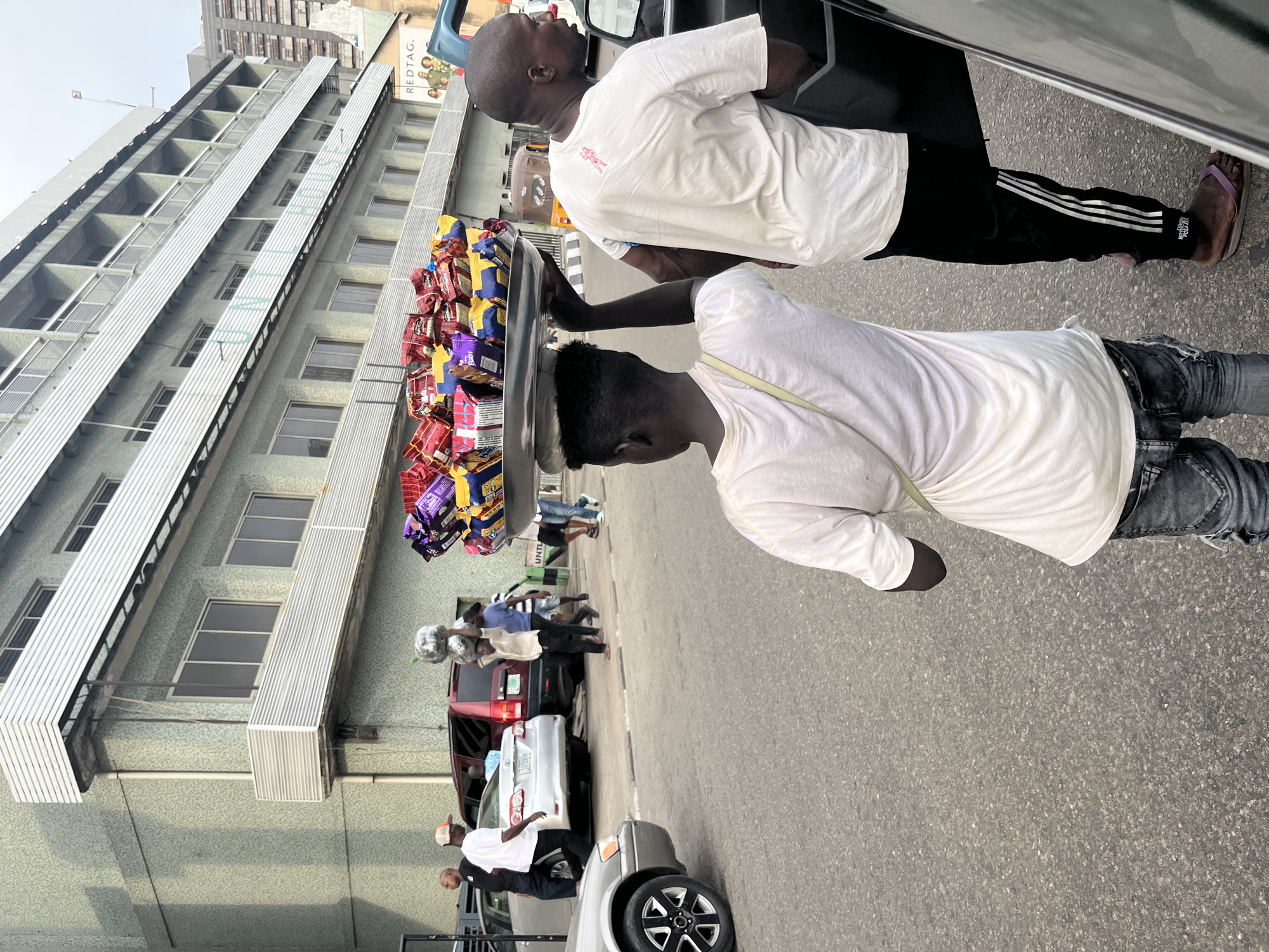 The image shows two men standing on the street next to cars. One man, wearing a white t-shirt and black and white striped pants, leans into a car window. The second man, wearing a white t-shirt and gray jeans, holds a tray filled with packaged snacks on his head. The scene takes place on a busy street in Lagos, Nigeria with cars, pedestrians, and a tall building in the background.