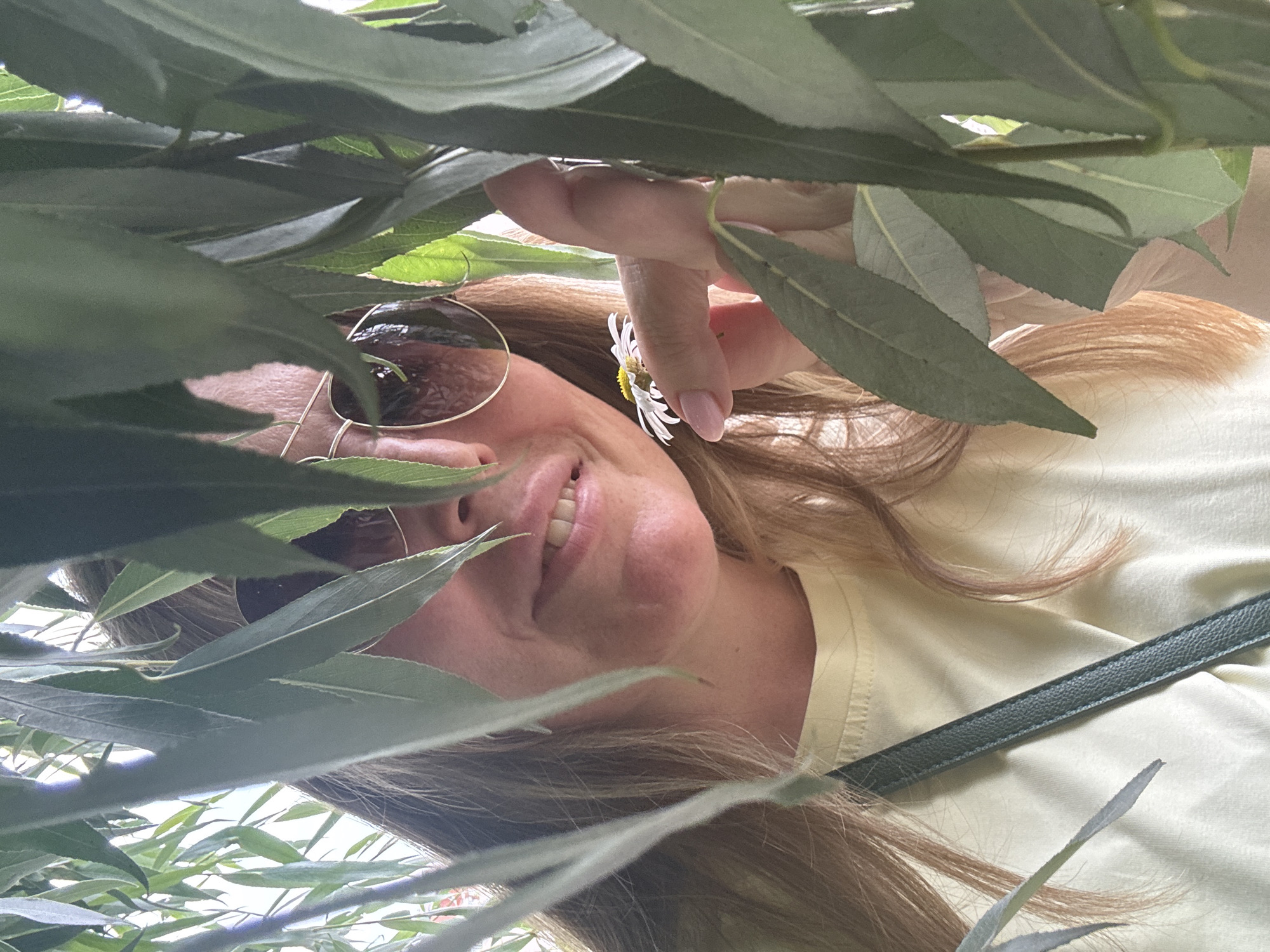 A blonde woman with sunglasses and a yellow t-shirt is partially obscured by green leaves, holding a small white flower in her right hand. She appears to be outdoors, possibly in a park or garden in Ufa, Russia, as indicated by the surrounding foliage and soft lighting. The woman is smiling and looking at the camera, creating a candid and natural portrait.