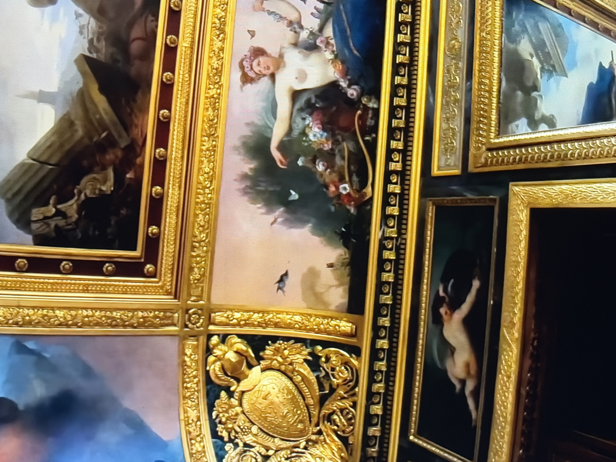 The image is a close-up, top-down view of a section of a ceiling adorned with multiple framed paintings and ornate gilded borders. The paintings depict classical and allegorical scenes.
In the upper right quadrant, a painting shows a fragmented classical ruin with a broken column lying on the ground, suggesting a historical or mythological setting.
To the left of the ruin painting, a smaller, darker rectangular painting features a gilded crest or coat of arms surrounded by elaborate gold foliage, set against a dark green background.
Below the crest and to the right of it, a larger horizontal painting depicts a nude woman with flowers in her hair, reclining amongst a profusion of flowers and greenery. Butterflies are visible in the scene, and she appears to be holding a floral garland.
Further down and centered, another horizontal painting shows a nude child or cherub in a supine position on a dark background, reaching out with one hand.
Below this, two more paintings are partially visible. The one on the left is a dark rectangle, and the one on the right is a larger rectangular painting with a gilded frame, showing a scene with clouds and what appears to be a flying chariot or winged figures.
The entire composition is framed by richly decorated gold borders featuring various patterns, including floral motifs, beaded elements, and intricate carvings. The colors are rich, with deep reds, blues, and golds dominating the decorative elements, and the paintings themselves rendered in muted, classical tones. The lighting suggests an indoor setting, likely a grand hall or palace, highlighting the opulent decoration.