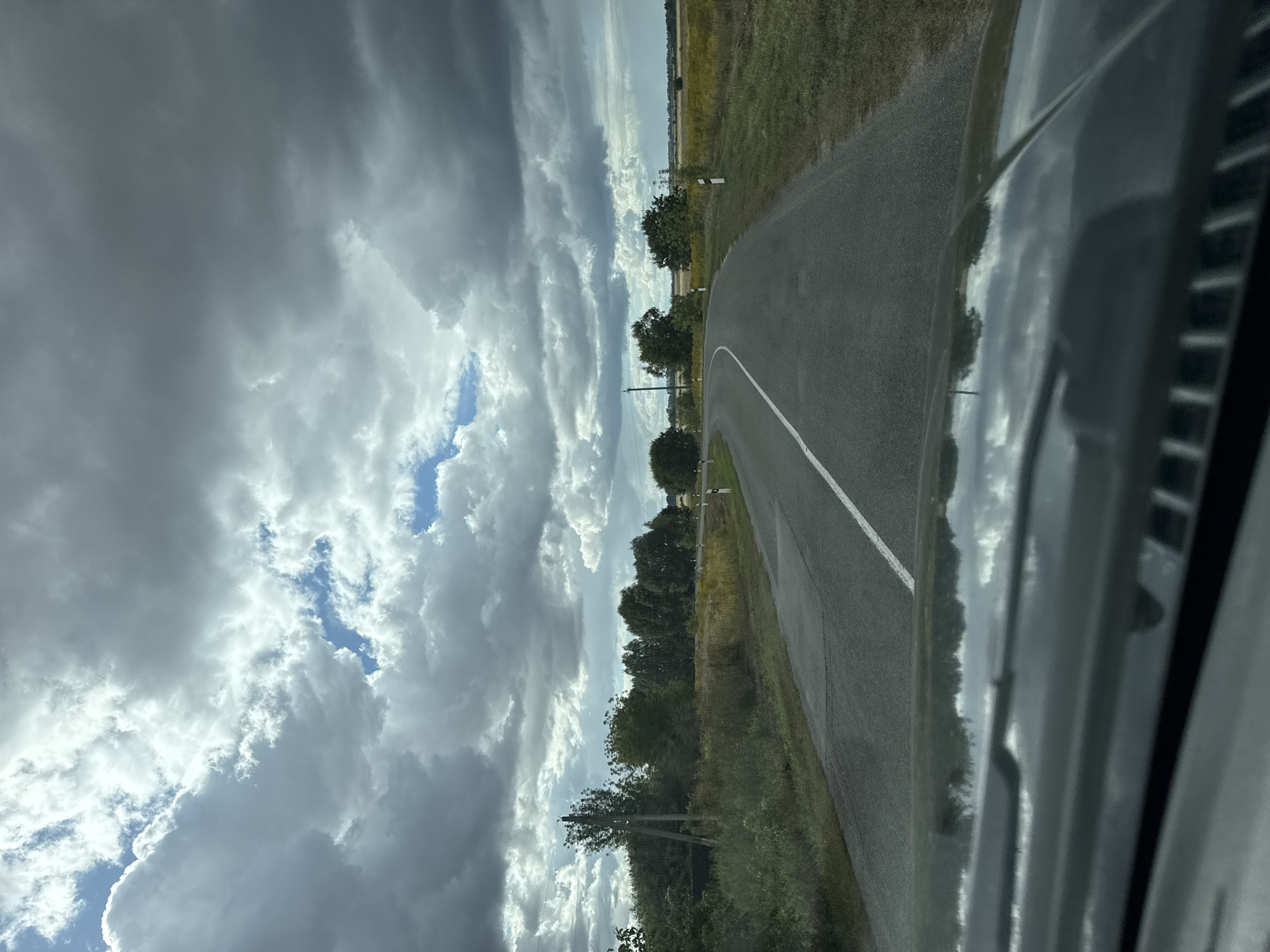 A slightly overcast, possibly approaching rainy, summer day is depicted from the perspective of someone inside a vehicle, looking out over the hood and windshield. The scene is a rural landscape featuring a gray asphalt road with a white center line that curves gently to the left. On either side of the road, there is green grass and vegetation, with trees and a field of yellow flowers visible in the distance. Tall, dark storm clouds dominate the sky, interspersed with patches of blue sky. A power pole is visible on the left side of the road. The reflection of the sky and trees can be seen on the car's hood. The location is identified as Fominka, Russia.