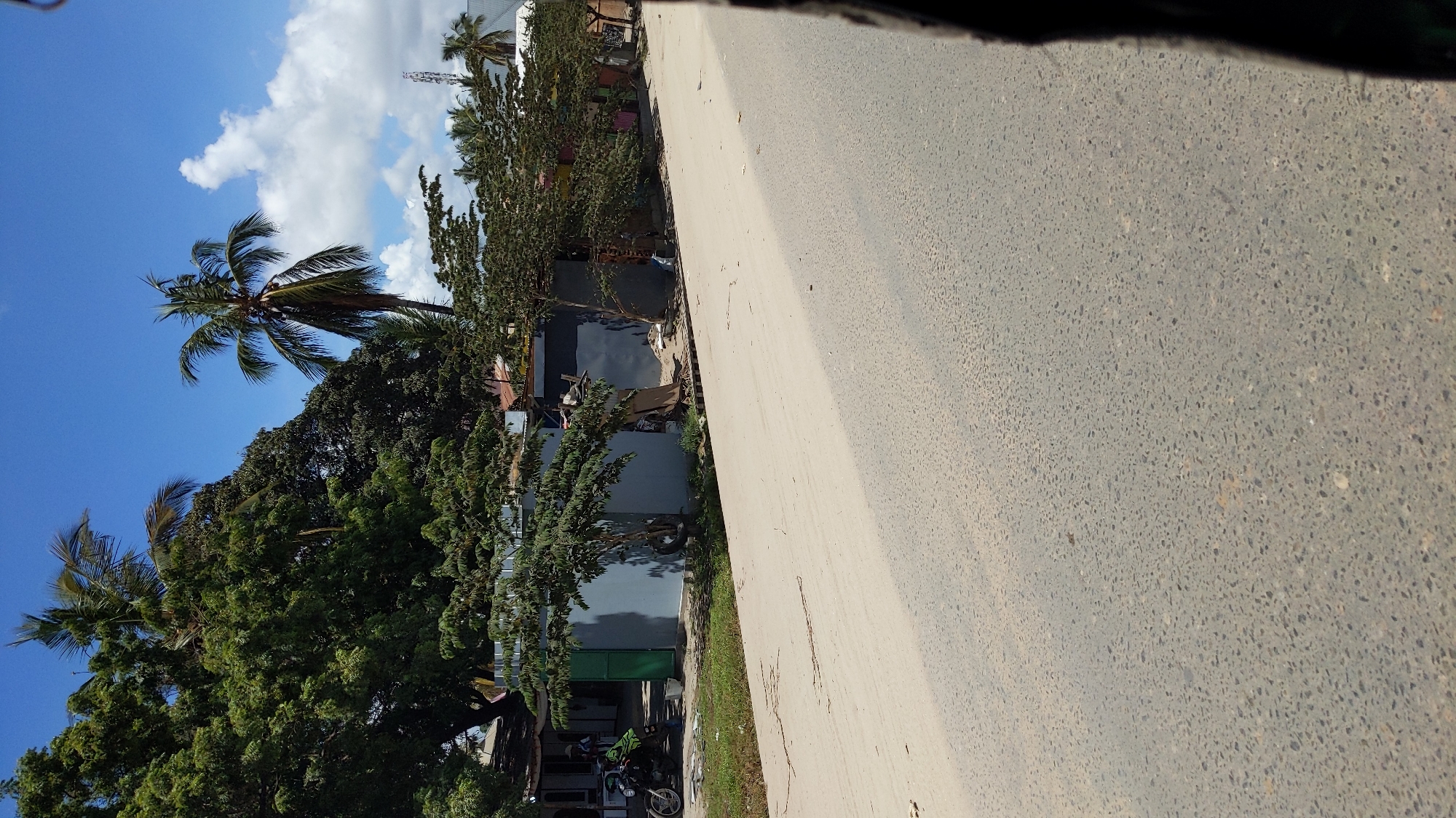 The image shows a street scene in Dar es-Salaam, Tanzania. A paved road takes up most of the foreground and right side of the image. Along the left side of the road are various buildings and trees, including palm trees. There are also motorcycles parked near one of the buildings. The sky is blue with scattered white clouds. The overall impression is of a sunny, tropical urban environment.