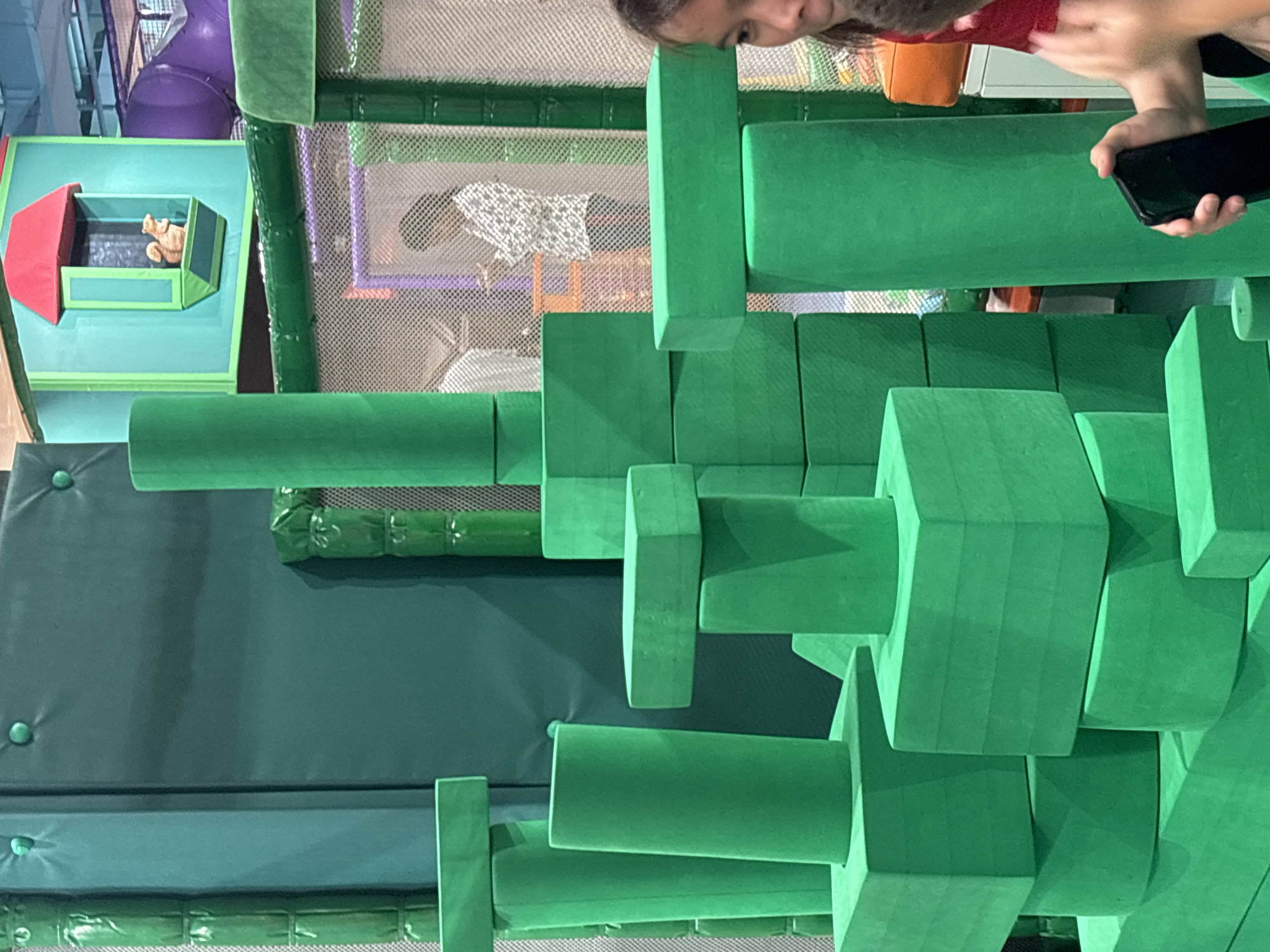 The image depicts a children's indoor play area, likely a playground or soft play center, with a prominent display of large, green, foam building blocks. The blocks are arranged in a structured, almost architectural manner, forming towers and platforms. In the background, a colorful play structure is visible, featuring a miniature house with a red roof and a window through which a small figurine, possibly a squirrel, is seen. Beyond this, a purple tube slide is partially visible. A person in a patterned shirt is seen in the mid-ground, looking towards a device. In the foreground, on the right side of the image, part of a person's face and shoulder are visible, holding a black smartphone. The overall environment appears to be a vibrant and engaging space for children. The exact location in Dubai, United Arab Emirates, cannot be determined from the visual information provided.