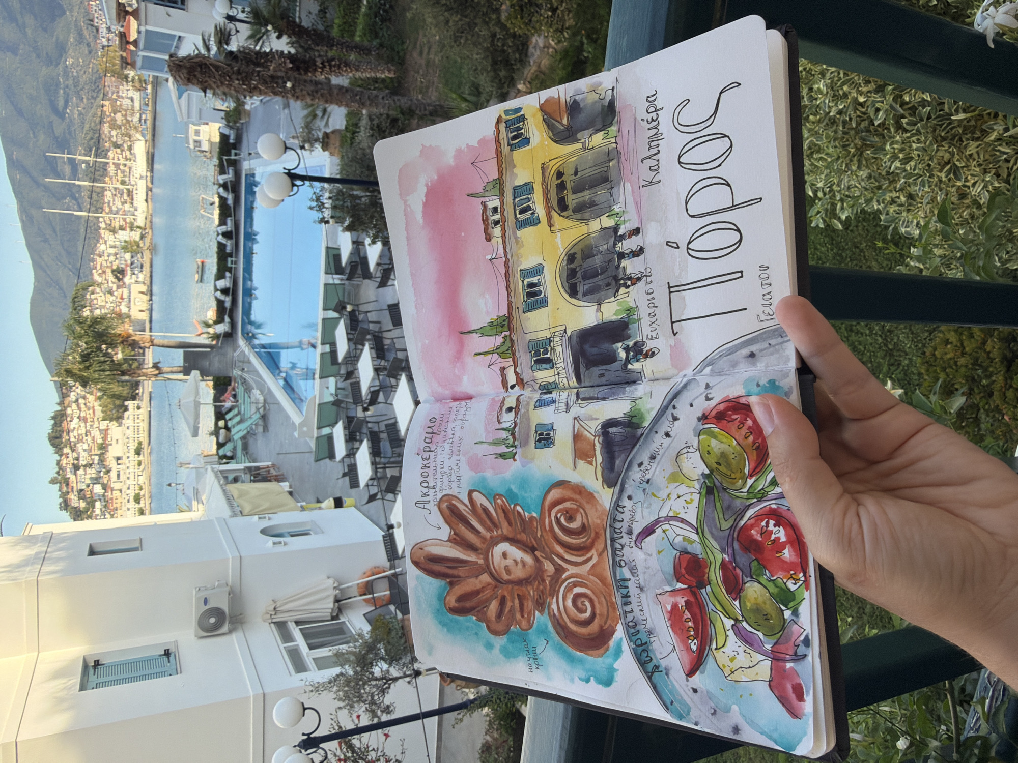 The image presents a first-person view of an open sketchbook held by a person, set against a vibrant outdoor background. The sketchbook, rendered in watercolor and ink, features several sketches.
On the right page, a depiction of a multi-story yellow building with blue shutters and dark roofs is seen. Above this sketch, the Greek word "Καλημέρα" (Good morning) is written. Below it, the name "Πόρος" (Poros) is prominently displayed in large letters, with "Γεια σου" (Hello/Goodbye) and "Ευχαριστώ" (Thank you) also visible.
The left page holds two distinct sketches. The upper sketch illustrates an "Ακροκέραμα" (Acrokerama), an architectural ceramic finial, possibly featuring a stylized figure. Accompanying Russian text describes it as the "keeper of the roof," featuring "human, flora figures" and "mythical images," found "on the corners of the roof." Below this, a colorful sketch of a "χωριάτικη σαλάτα" (Horiatiki Salata), or Greek salad, filled with tomatoes, olives, onions, and feta cheese, is shown. Russian text translates this as "Greek salad - literal translation, village salad."
A hand holds the bottom right corner of the sketchbook. The background reveals a sunny, clear day over a scenic coastal town, identified as Poros, Greece. White buildings, some with air conditioning units, palm trees, and an outdoor swimming pool area with numerous tables and chairs are visible in the foreground. Beyond this, the tranquil sea is dotted with boats, and a distant town with white houses climbs up a verdant mountainside, under a bright blue sky. The scene evokes a pleasant and artistic travel experience.
