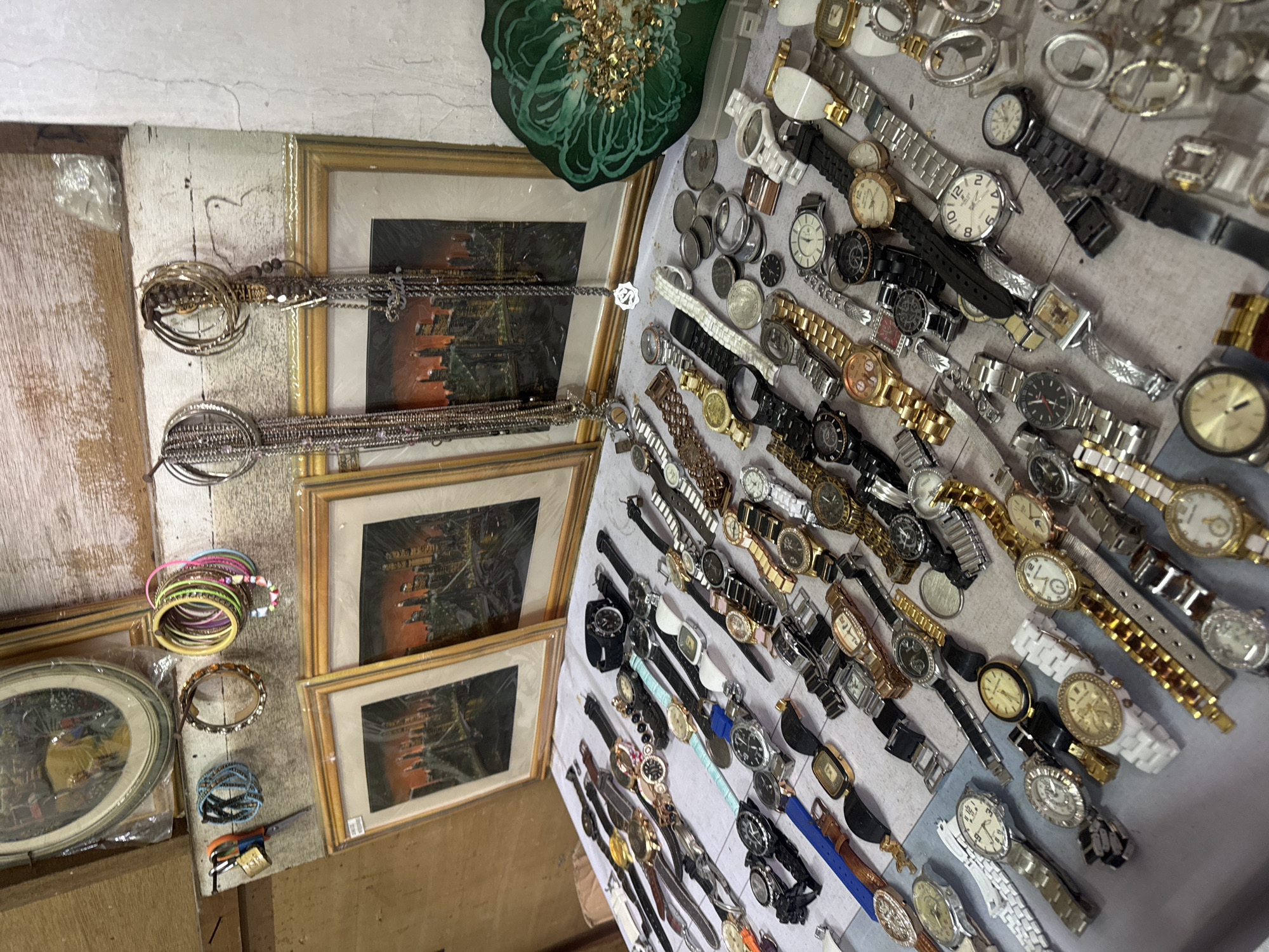 The image depicts a street market stall in Denpasar, Indonesia, likely selling watches and various accessories. The stall features a wide assortment of wristwatches, neatly arranged on a table. The watches vary in style, material, and color, with many having metallic bands in silver, gold, and black, as well as some with leather or fabric straps in blue and white. Some watches have Roman numerals on their faces, while others have standard numerical markers or are purely decorative.
In addition to watches, several other items are visible. To the left of the watches, there are framed pictures depicting cityscapes, possibly of New York. Above the framed pictures, a collection of colorful bracelets and bangles are displayed, along with a padlock and scissors. Hanging from hooks above the stall are long, decorative necklaces or chains. A distinct green, round decorative item with gold flakes is positioned on the right side of the stall. The overall environment suggests an outdoor market setting with a rustic wooden backdrop and a textured white wall. The lighting indicates it is daytime.