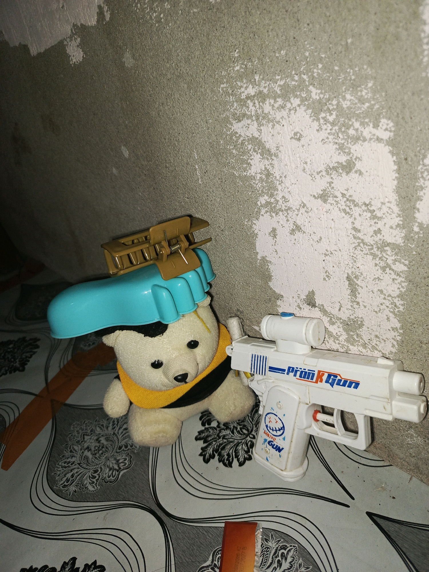 An indoor scene depicts a plush teddy bear wearing a yellow scarf and a blue plastic object on its head, topped with a brown hair clip. Next to the teddy bear is a white plastic toy gun with blue lettering that reads "Pran R Gun". The subjects are positioned on a patterned floor against a textured wall with peeling paint. The setting is a room within a house, potentially in Megang Sakti I, Indonesia.