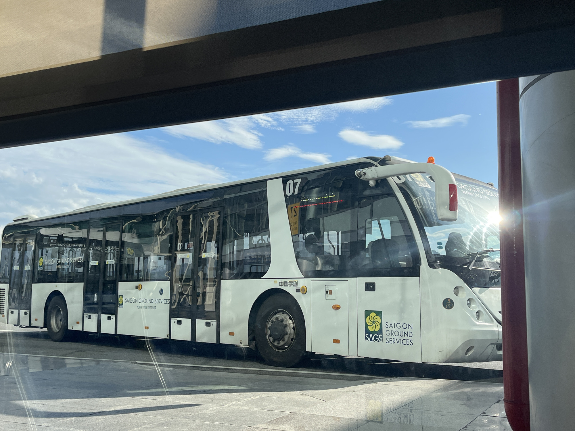 A long white airport shuttle bus, with black windows and lower trim, is parked on an asphalt or paved surface under a bright blue sky with scattered cirrocumulus clouds. The bus displays "SAIGON GROUND SERVICES" and "SAGS" branding in green and white on its sides, along with the slogan "YOUR TRUE PARTNER." The bus number "07" is prominently displayed on its front-side. Near the rear wheel well, Chinese characters "中国宇通" (Zhōngguó Yǔtōng), indicating the manufacturer Yutong, are visible. A handicap accessibility symbol is present on the front of the bus. Reflections of overhead structures and internal elements are visible on the bus windows and the ground. The image is taken through a window, with a dark horizontal frame partially obstructing the top. Intense sunlight from the right creates a strong lens flare near the bus's front windshield and a vertical red and grey pole. This scene is located at Đà Nẵng International Airport, Đà Nẵng, Vietnam.