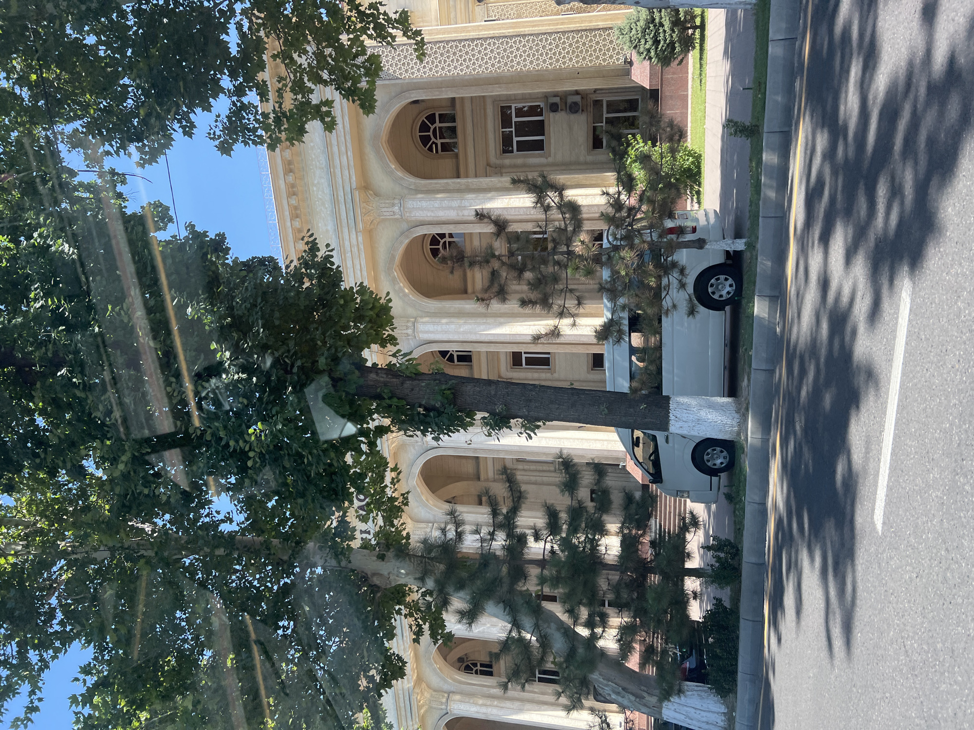 The image shows a white minibus parked on the side of a street in Tashkent, Uzbekistan, in front of a large, ornate building. The building has a classical architectural style with arched windows and decorative columns. Trees with lush green foliage line the street, casting shadows on the asphalt road. The sky is clear and blue, indicating a sunny day. The scene appears to be a public space, possibly a government building or a cultural institution, given the grandeur of the architecture and the presence of the minibus, which might be used for transportation of staff or visitors.