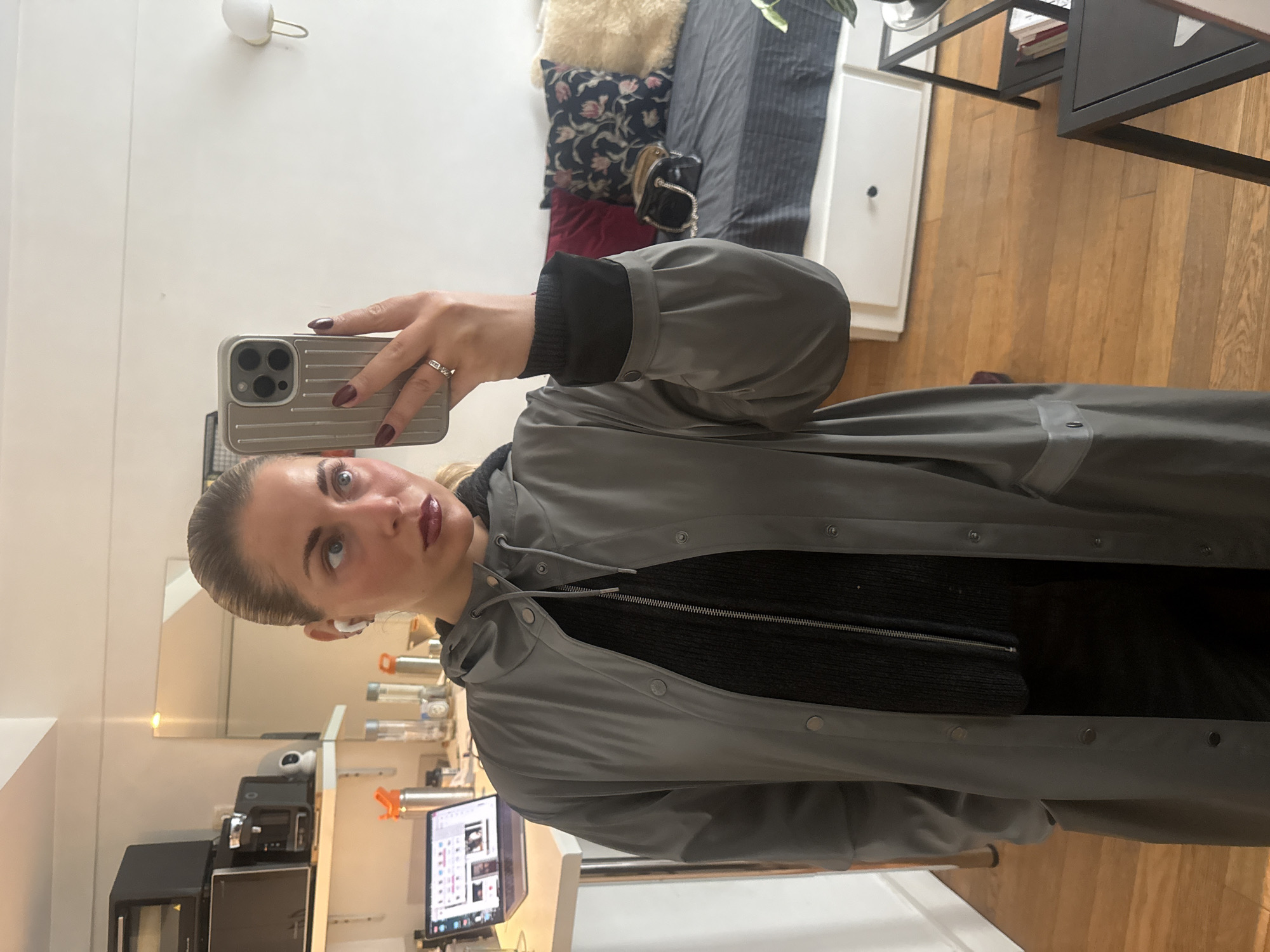 A fair-skinned woman is taking a selfie in what appears to be an indoor setting. She is wearing a grey rain jacket and a black zip-up sweater. An earbud is visible in her ear. The room has a wooden floor. In the background are some shelves and what appear to be kitchen appliances, including a coffee maker and a microwave. A computer monitor sits on a desk. The scene does not provide sufficient details to confirm the location in the city of Paris, France.