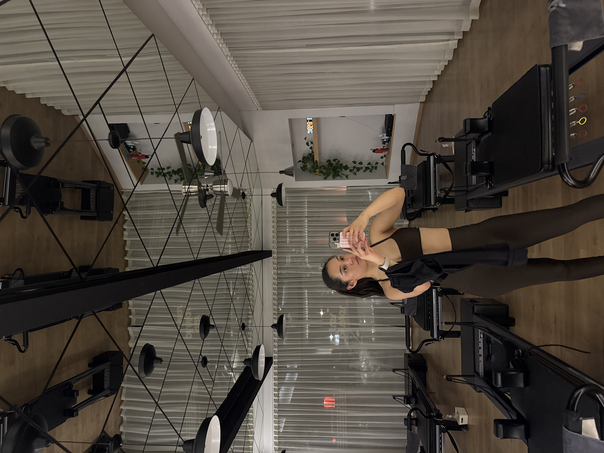 A woman is standing in a workout studio, facing the camera, taking a photo of herself in a mirror. The studio contains pilates reformers. The studio has white blinds, a wooden floor, and a ceiling with exposed lights. There is a mirror running along the wall in front of her. Puebla, Mexico is not verifiable from the image.