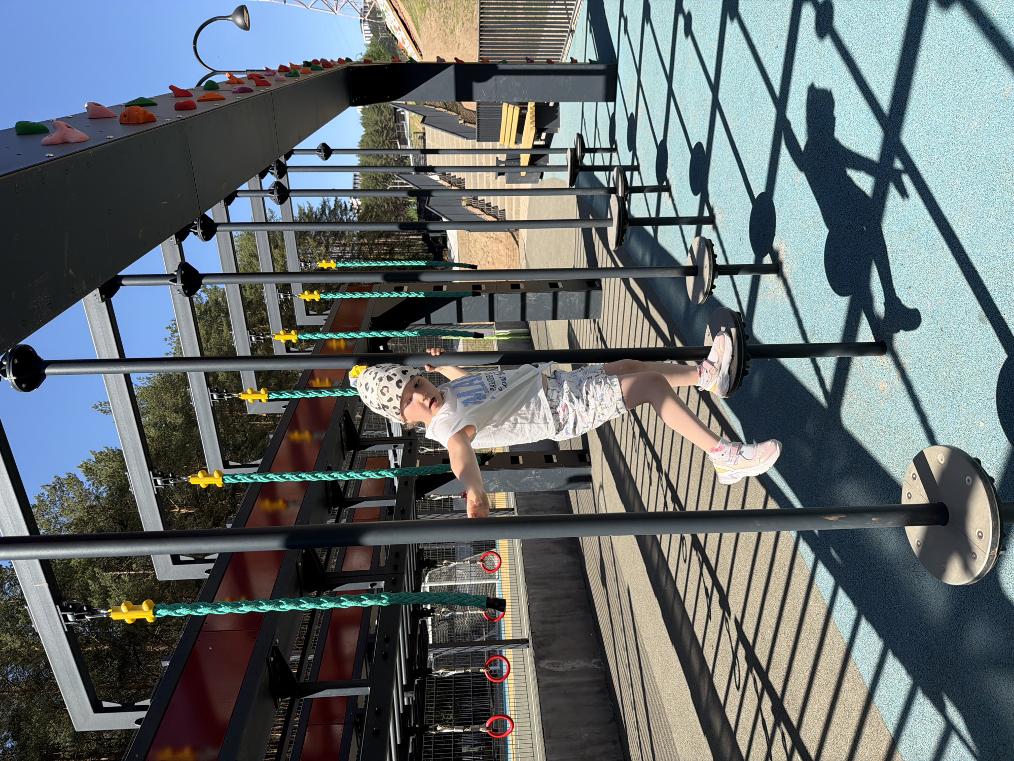 A child is hanging on playground equipment, seemingly at a park in Izhevsk, Russia. The equipment consists of parallel horizontal bars with attached climbing ropes. A climbing wall is also visible. The child, wearing light-colored clothing and a patterned hat, appears to be suspended between the bars. Sunlight casts shadows of the equipment onto a blue surface beneath.