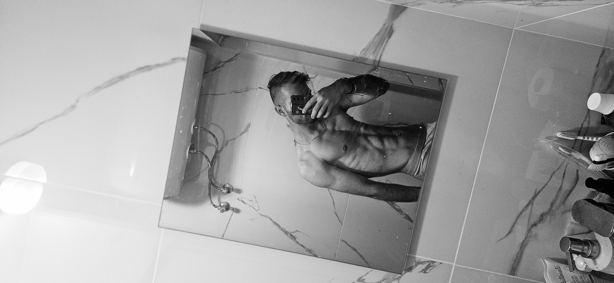 The image is a black and white photo taken in a bathroom, likely as a mirror selfie. The subject is a shirtless man with visible abdominal muscles and a necklace. He is holding a smartphone, presumably to capture the photo. The bathroom features marble-patterned tiles, a sink with visible plumbing, and a countertop with personal care items like toothbrushes, a cup, and various bottles. There's no information in the image to suggest the exact location is in Plovdiv, Bulgaria.