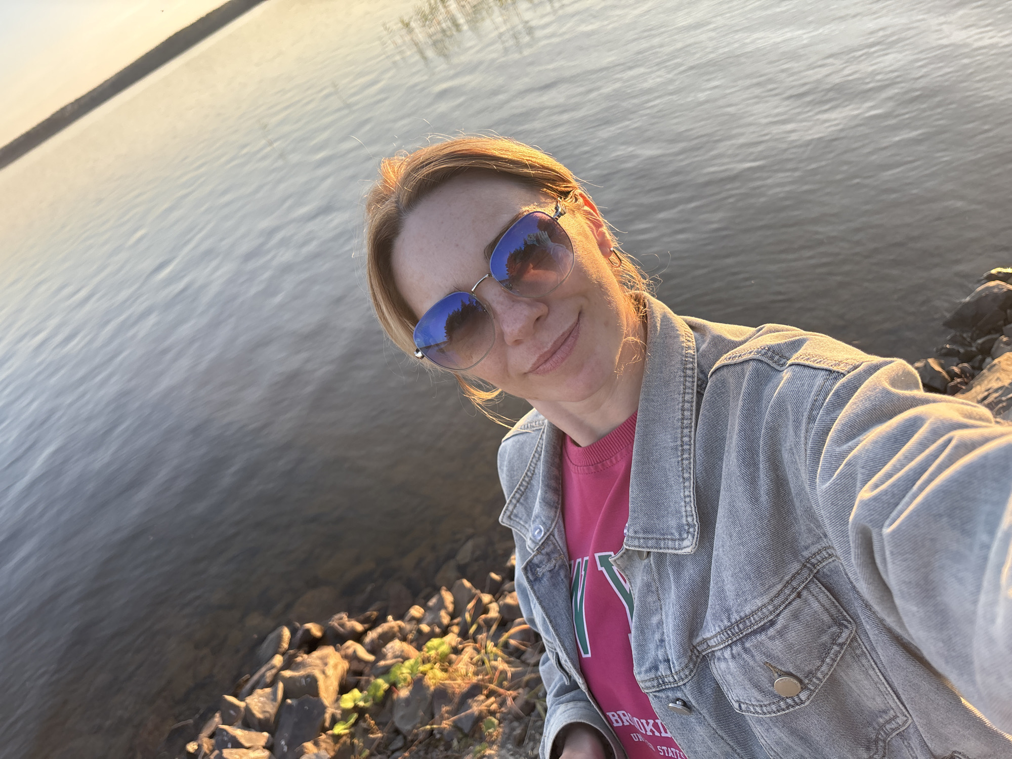 A woman, appearing to be in her 30s, with blonde hair and wearing sunglasses, a pink t-shirt, and a denim jacket, takes a selfie while standing by a body of water. The environment appears to be a natural setting, with rocks and vegetation visible along the shoreline. The water is calm and reflects the light of what appears to be either sunrise or sunset. The overall impression is of a casual, outdoor moment. The image was taken in Shuiskaya Chupa, Russia.