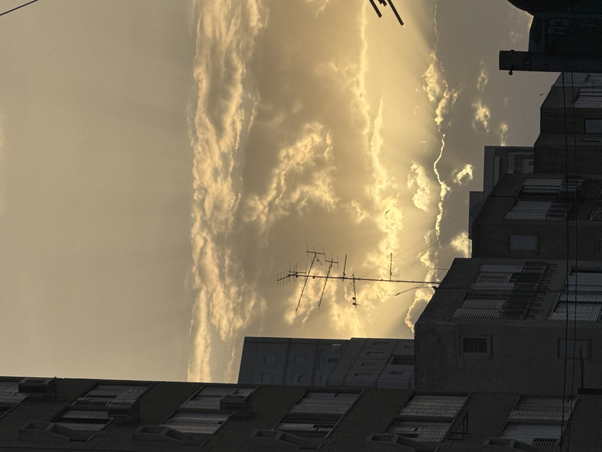 The image depicts a view in Ramat Gan, Israel, likely at dusk or dawn, with buildings silhouetted against a bright, cloud-filled sky. An antenna stands prominently on the roof of one of the buildings. The sky is filled with golden light, creating a striking contrast with the dark buildings. The buildings are typical of urban architecture, with rectangular shapes and rows of windows. No people are visible. The presence of an antenna suggests residential use.