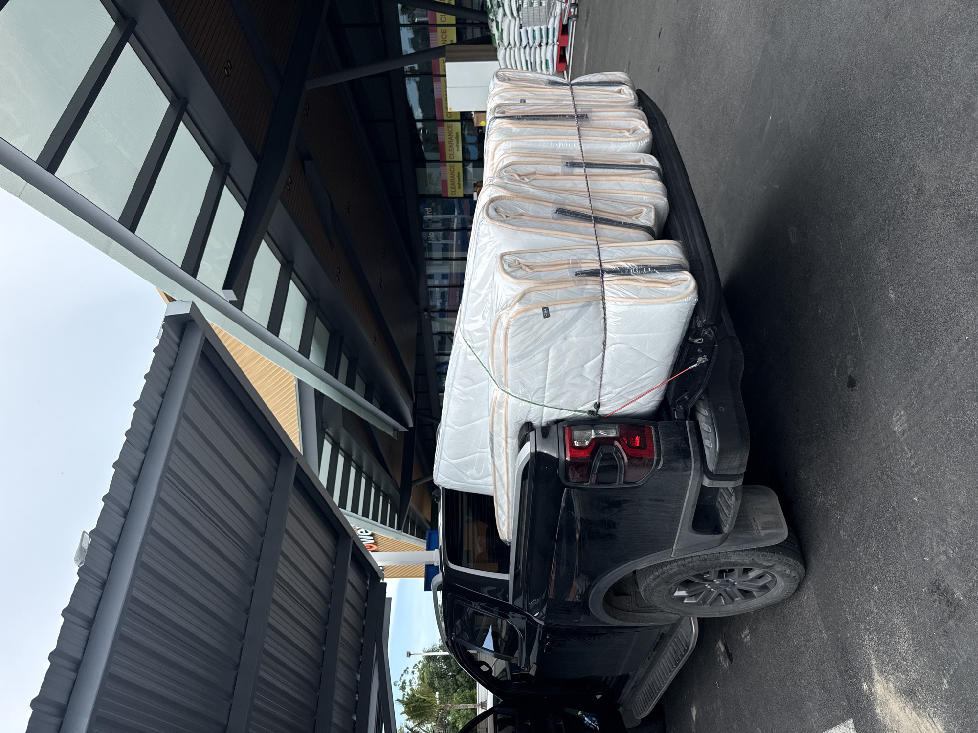 A black pickup truck is parked in what appears to be a parking lot with a covered structure overhead. The truck bed is loaded with several mattresses, all wrapped in clear plastic and tied down with ropes. The mattresses are stacked tightly and extend slightly over the sides of the truck bed. The background shows a building with large windows and signage that includes the word "CLEARANCE." There are also stacks of white bags and shopping carts visible in the background. The ground is paved asphalt. The scene is likely in Baan Chaweng Noi, Thailand, given the context.