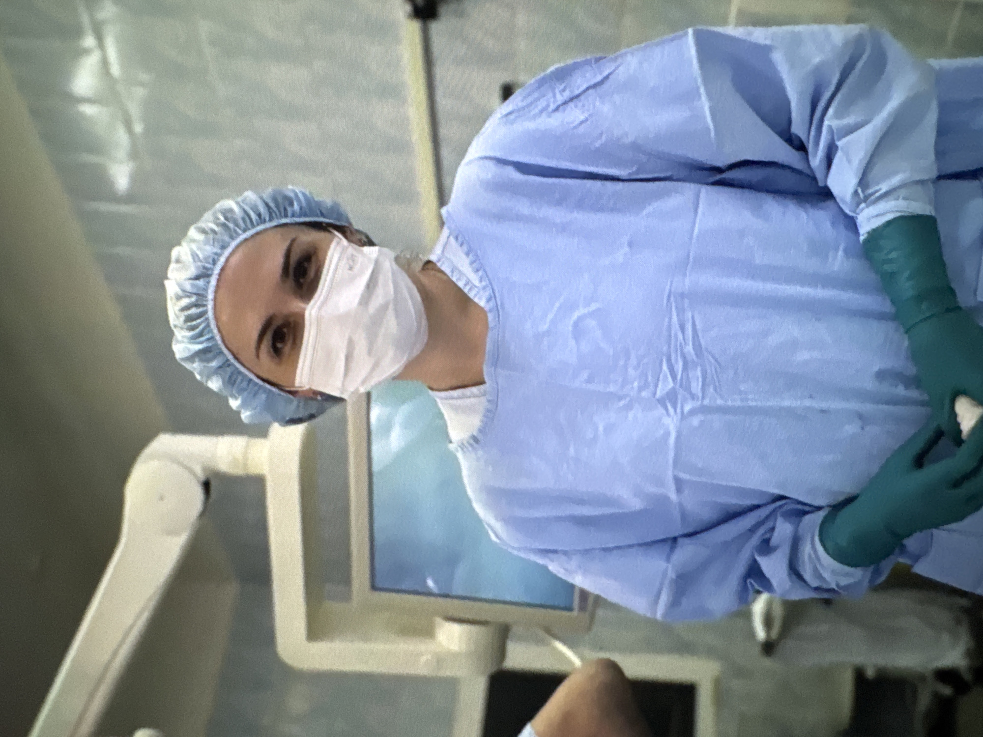 The image displays a female medical professional within a sterile medical facility, likely an operating room, in Sertolovo, Russia. She is centrally positioned, directly facing the viewer. Her attire includes a light blue surgical scrub cap, a white surgical mask obscuring her nose and mouth, a light blue long-sleeved surgical gown, and dark green surgical gloves. Her eyes are visible above the mask, maintaining direct visual contact. Her gloved hands are clasped at waist level, holding a small, light-colored object, consistent with medical gauze or cotton.
The background depicts a clean, brightly lit clinical environment. To the left, a large, articulated white medical equipment arm extends from the ceiling. Below it, a monitor displays an indistinct light blue and white image. The walls appear to be light blue-grey, possibly panelled. A partial view of another individual's arm and shoulder is visible on the far left. The scene suggests a professional medical setting.